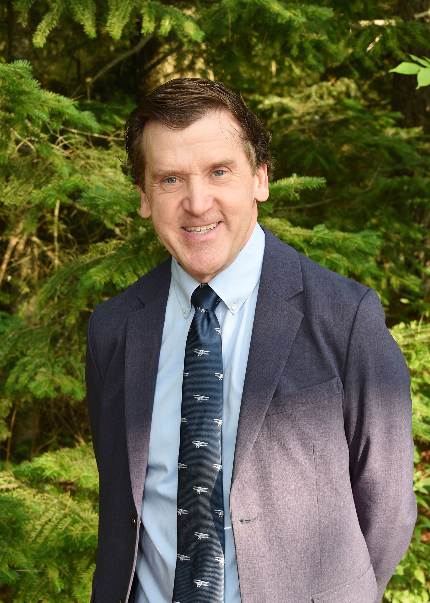 Man in a navy blazer and patterned tie smiles against a background of green trees.