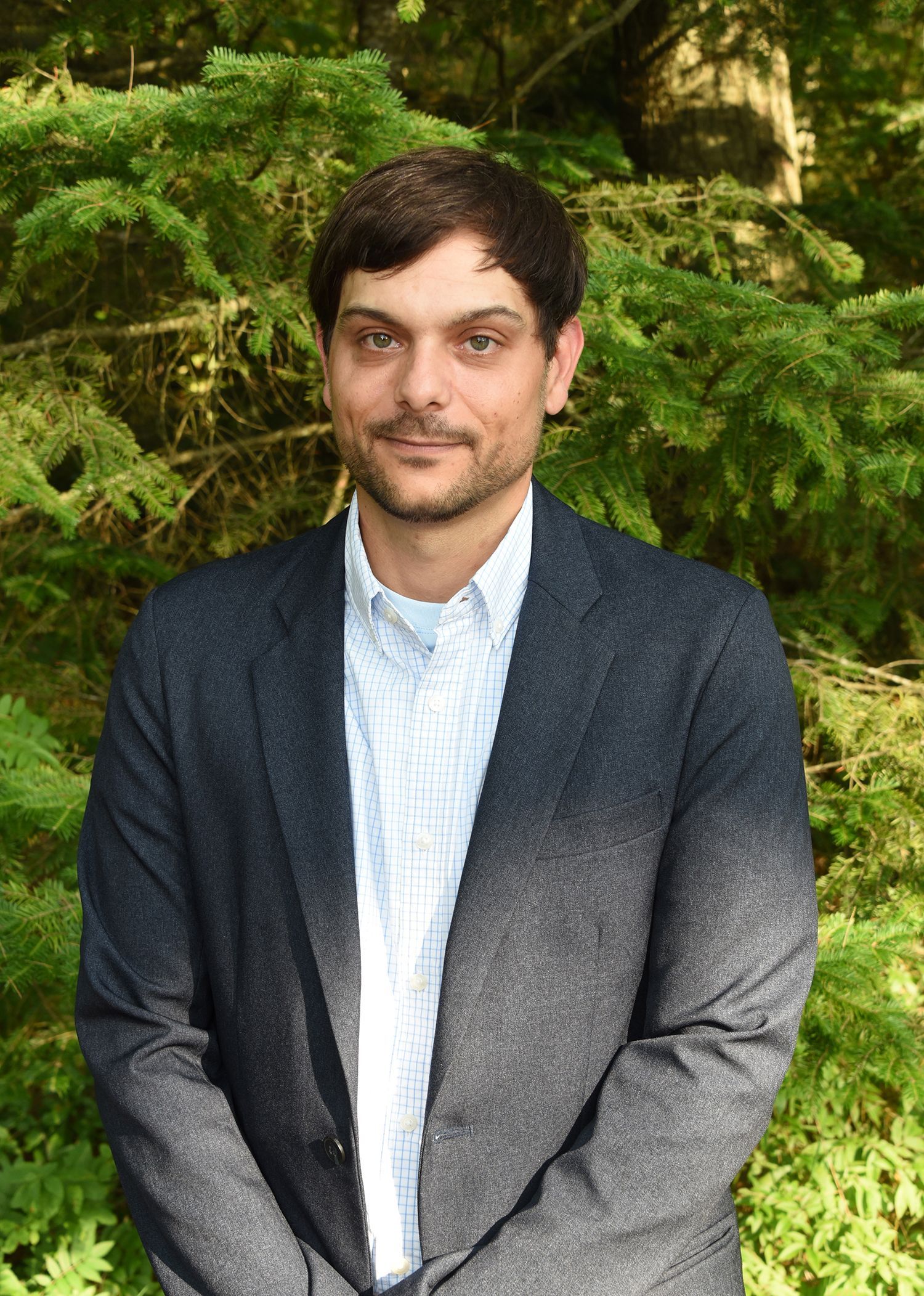 Man in a dark blazer and blue shirt smiling, standing outside in front of lush green foliage.