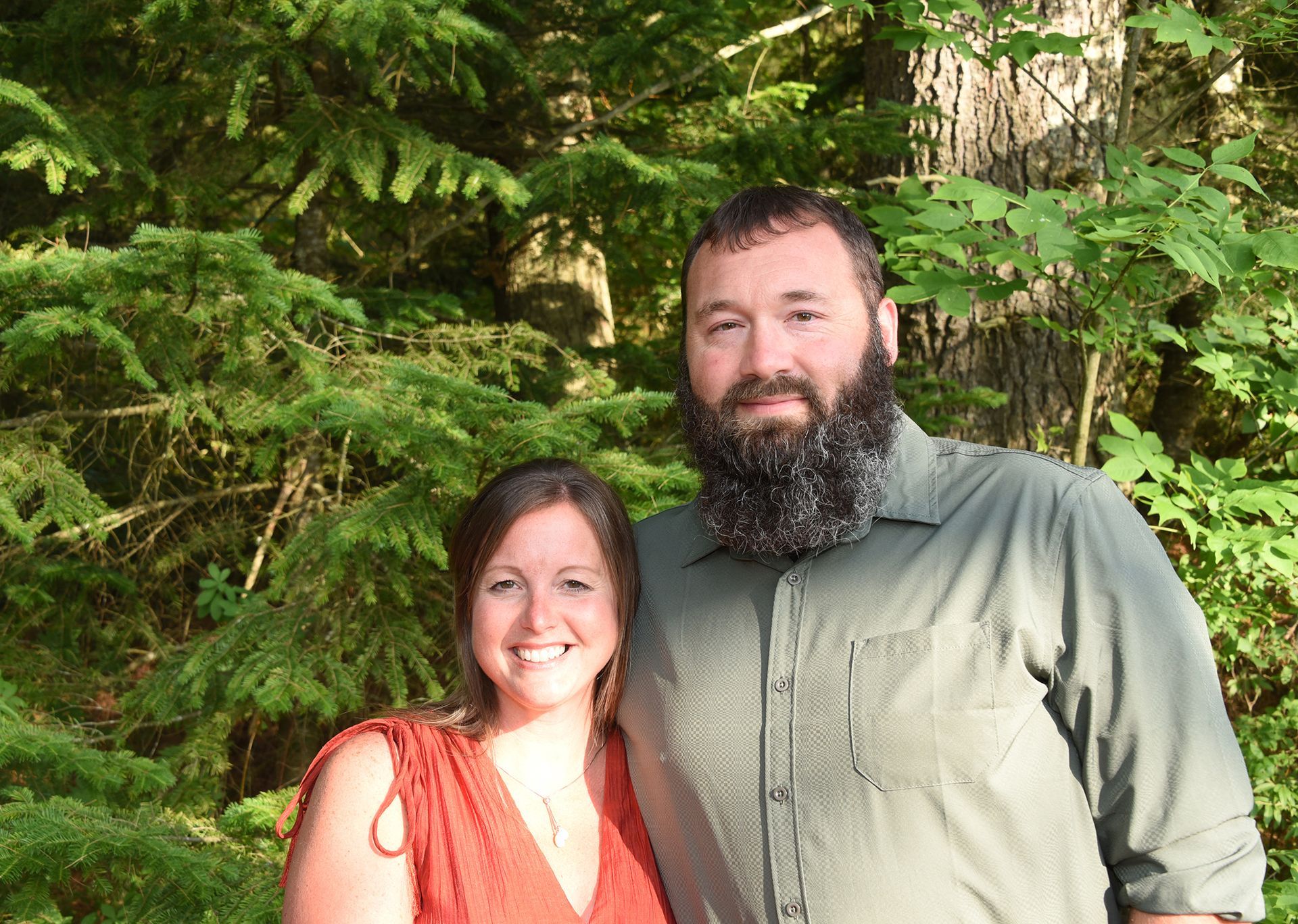Woman in red dress and bearded man in green shirt smiling in front of trees.