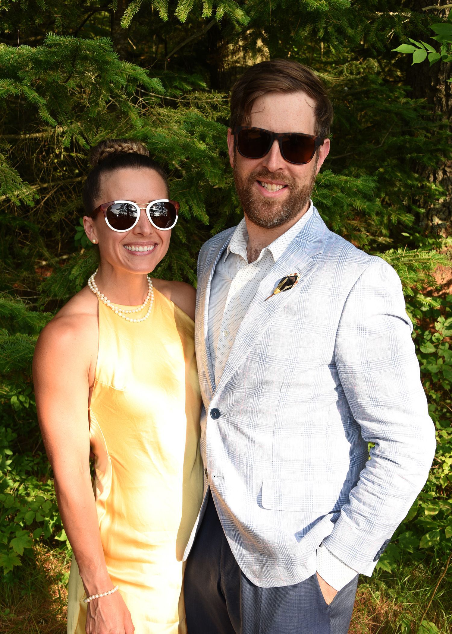 A smiling couple poses outdoors. Woman in a yellow dress and sunglasses, man in a light blue blazer, both wearing sunglasses.