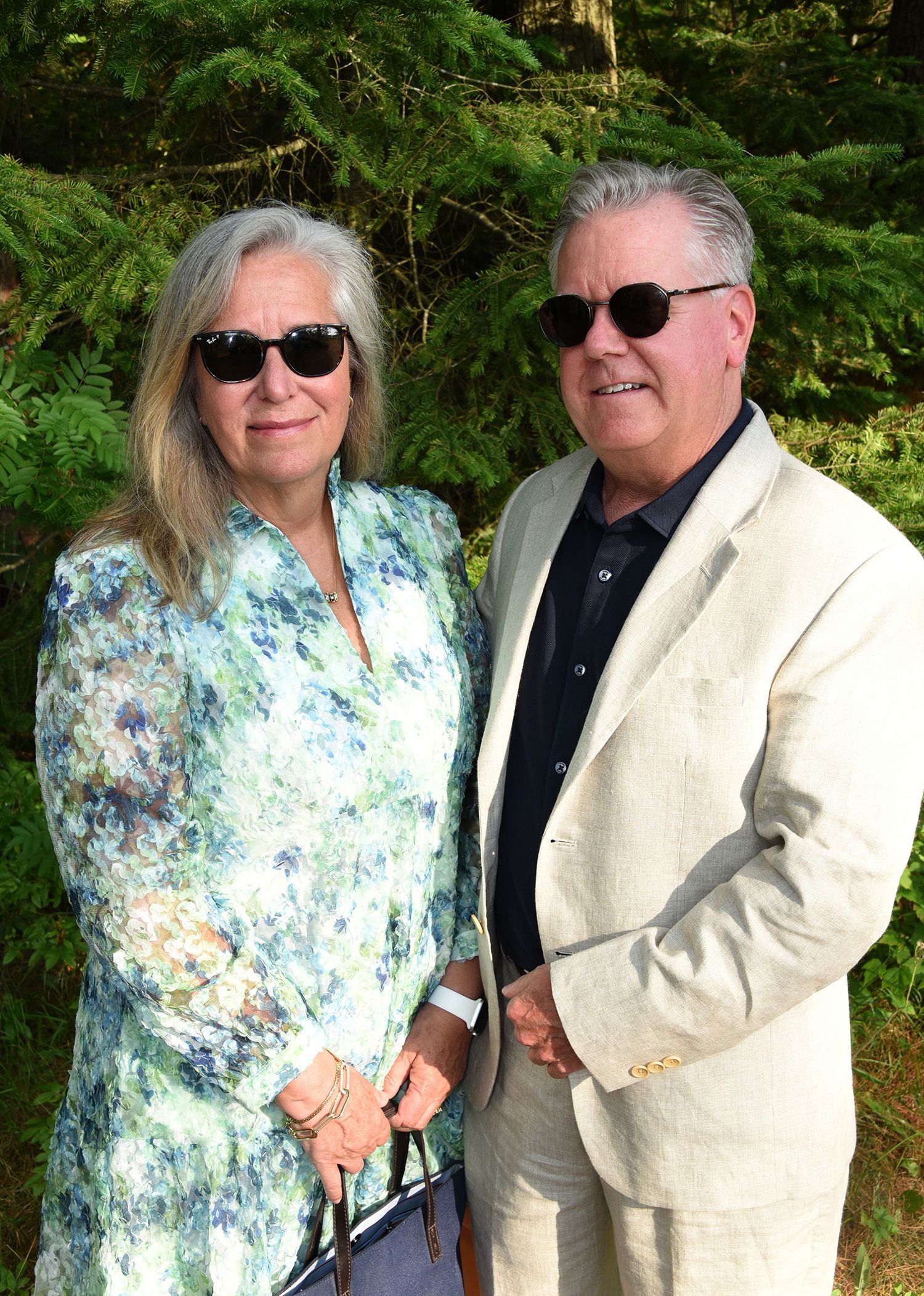 A man and woman in sunglasses pose together outdoors. The woman wears a patterned blouse; the man, a light-colored jacket.