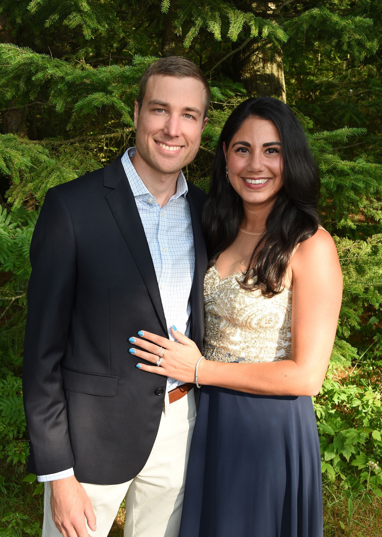 A smiling couple poses outdoors; man in a blazer, woman in a strapless gown, her engagement ring visible.