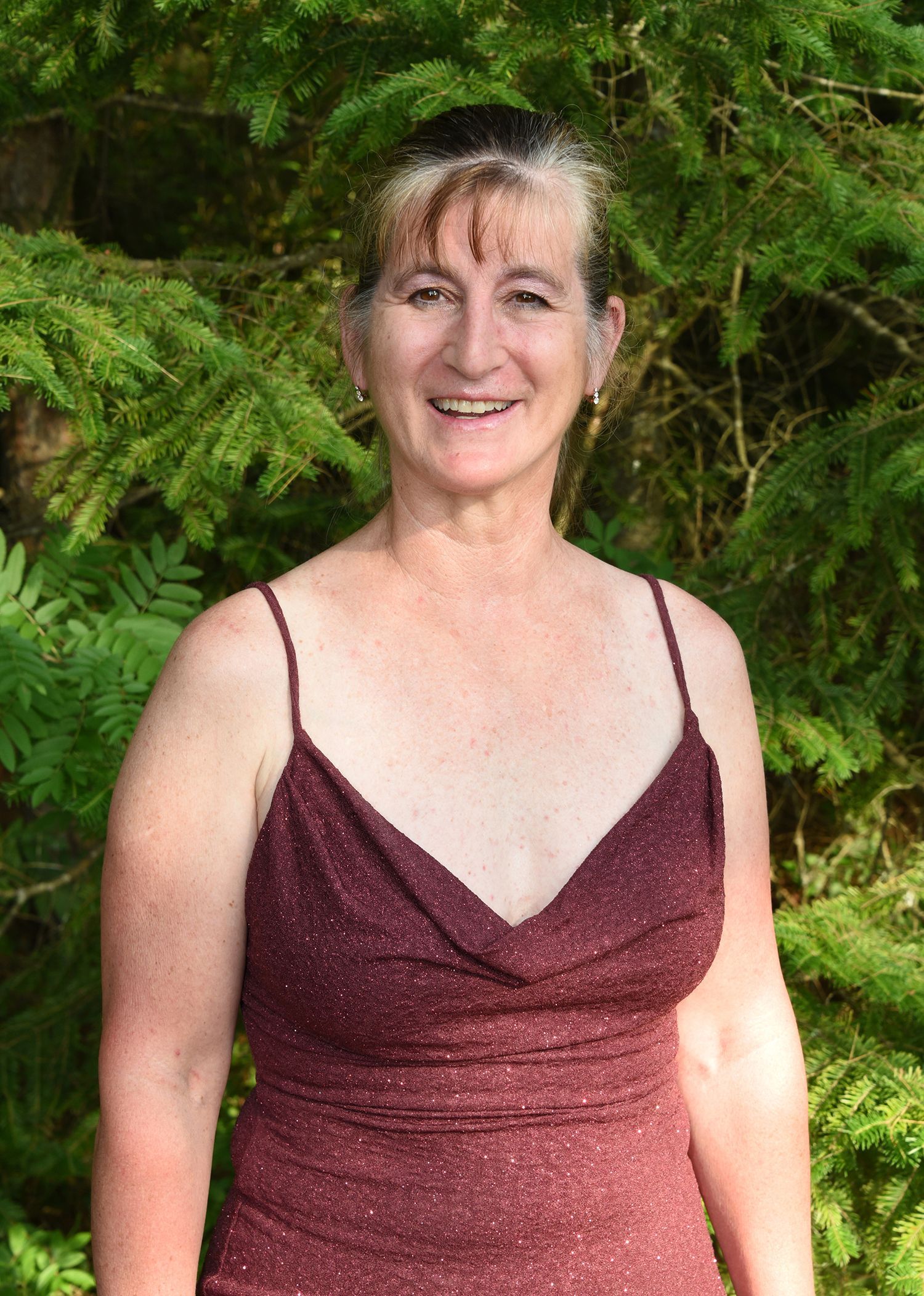 Woman smiling in a burgundy dress with spaghetti straps, standing in front of dark green foliage.