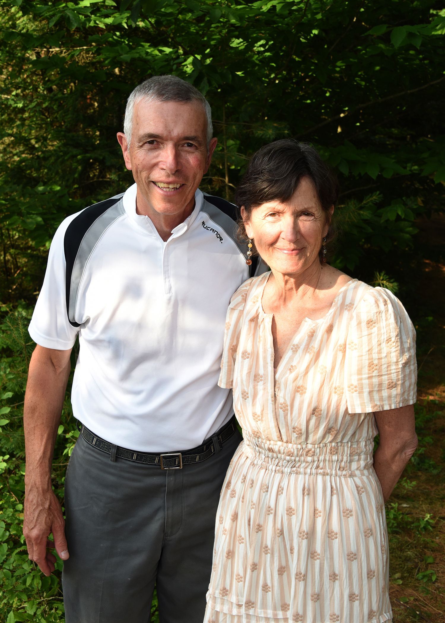 Older couple standing together in front of trees. Man wears a white polo shirt and grey pants, woman wears a patterned dress.