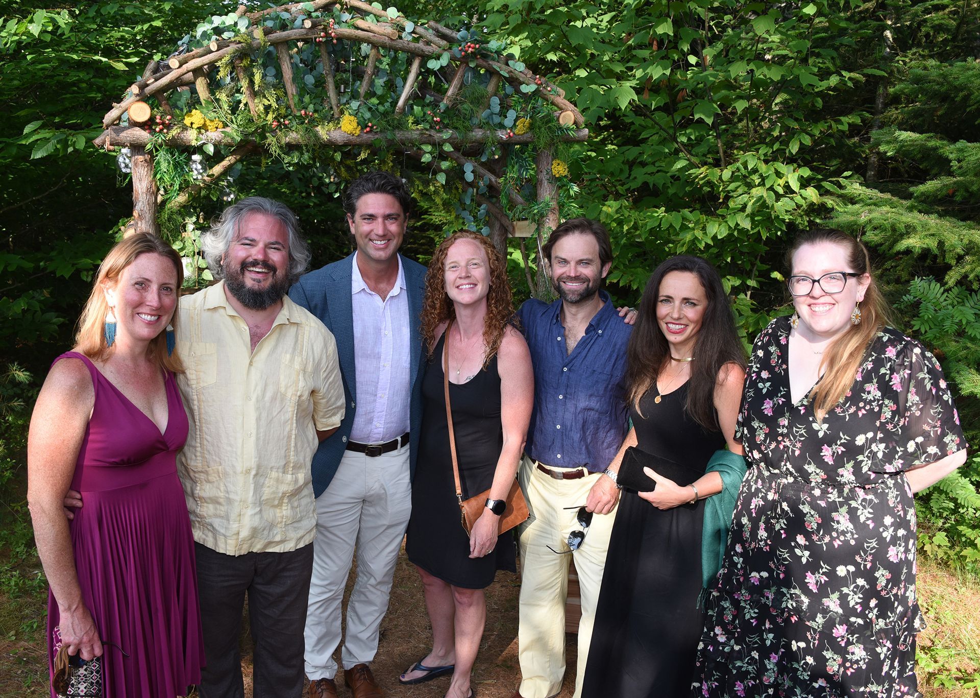 Group of seven people smiling for a photo under a wooden archway decorated with foliage, in a lush, green outdoor setting.