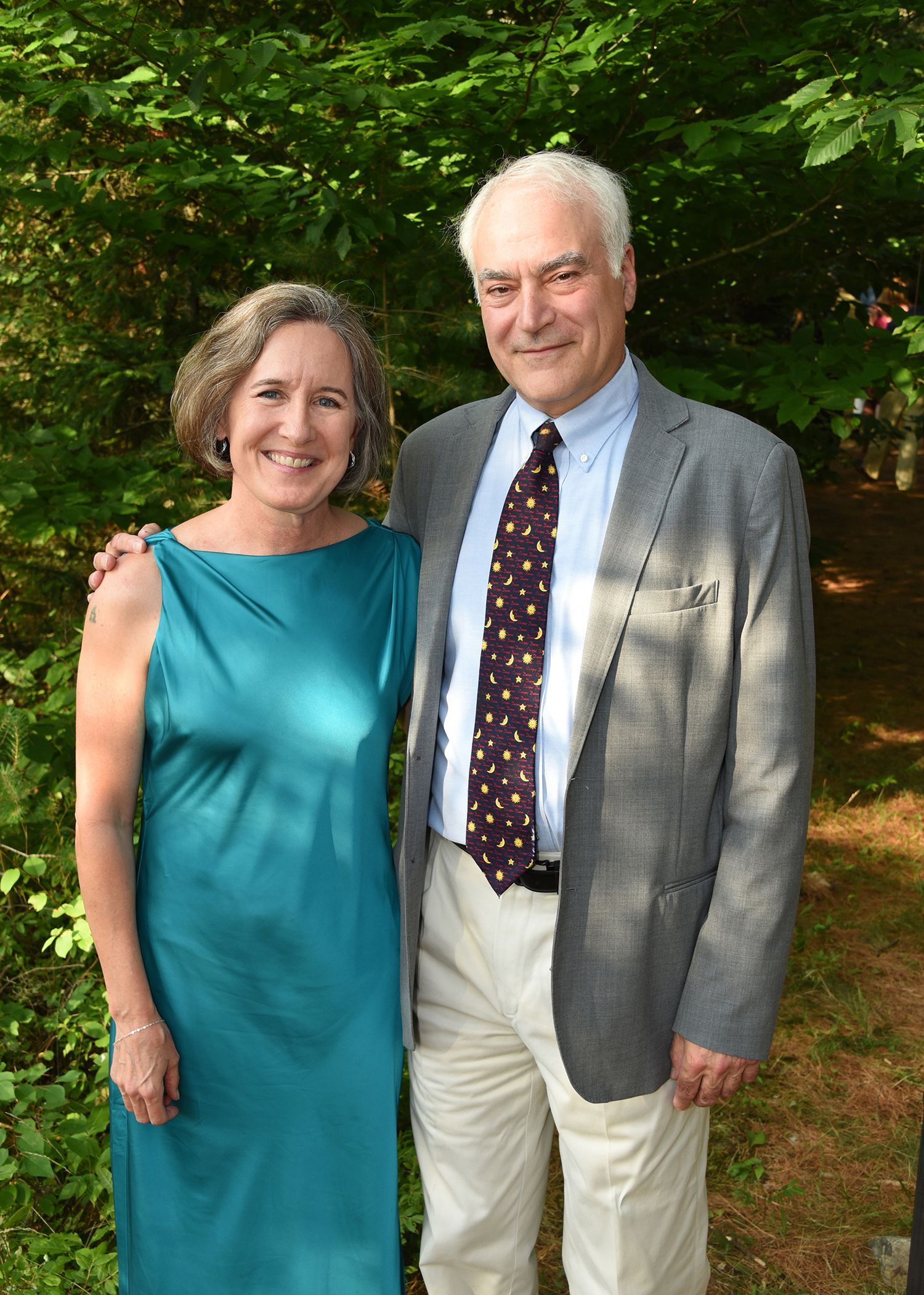 An older couple stands together outdoors; the woman wears a teal dress, and the man wears a jacket, tie, and khakis.