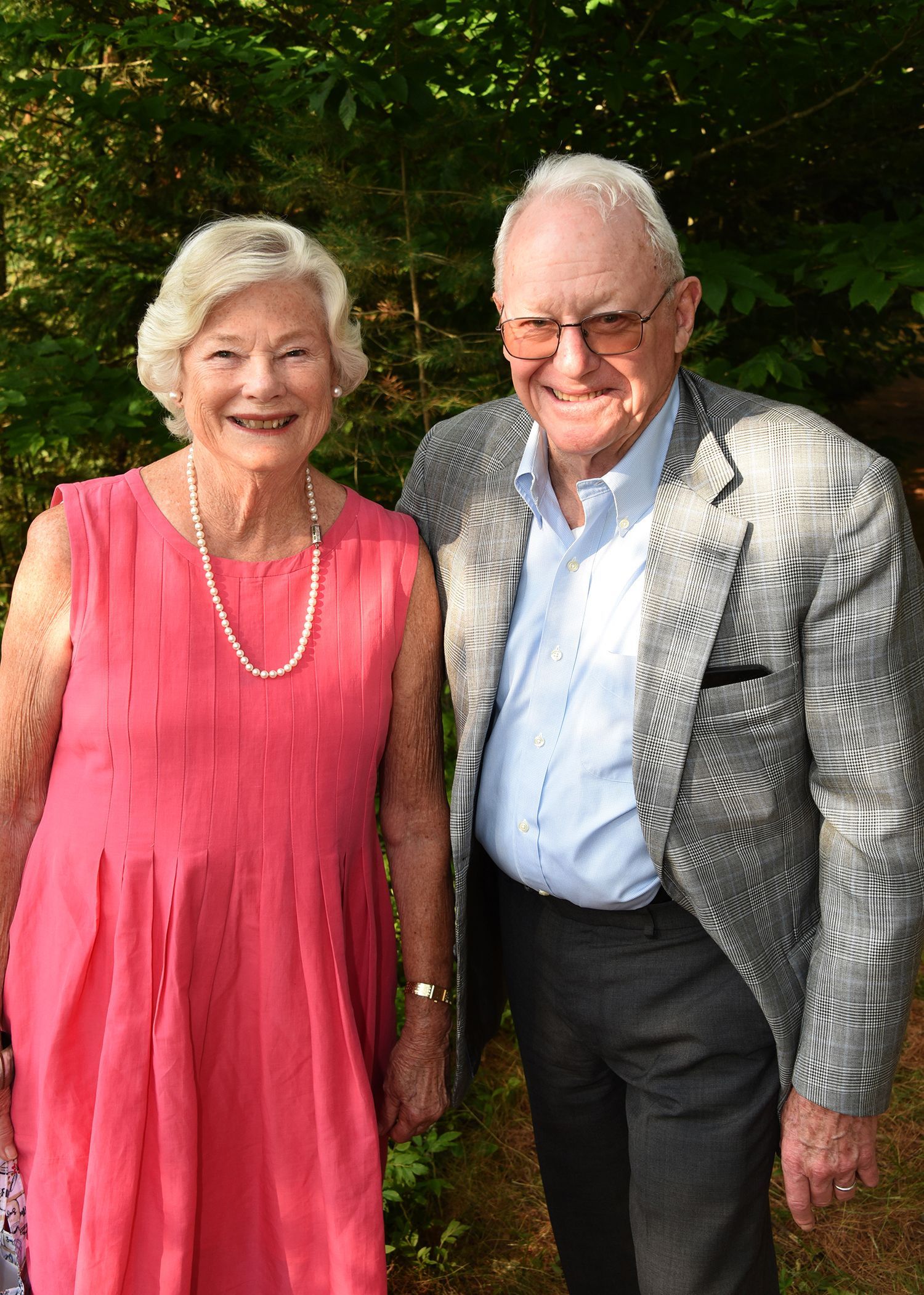 An elderly couple stands side-by-side outdoors. The woman wears a pink dress and necklace, the man a patterned jacket.