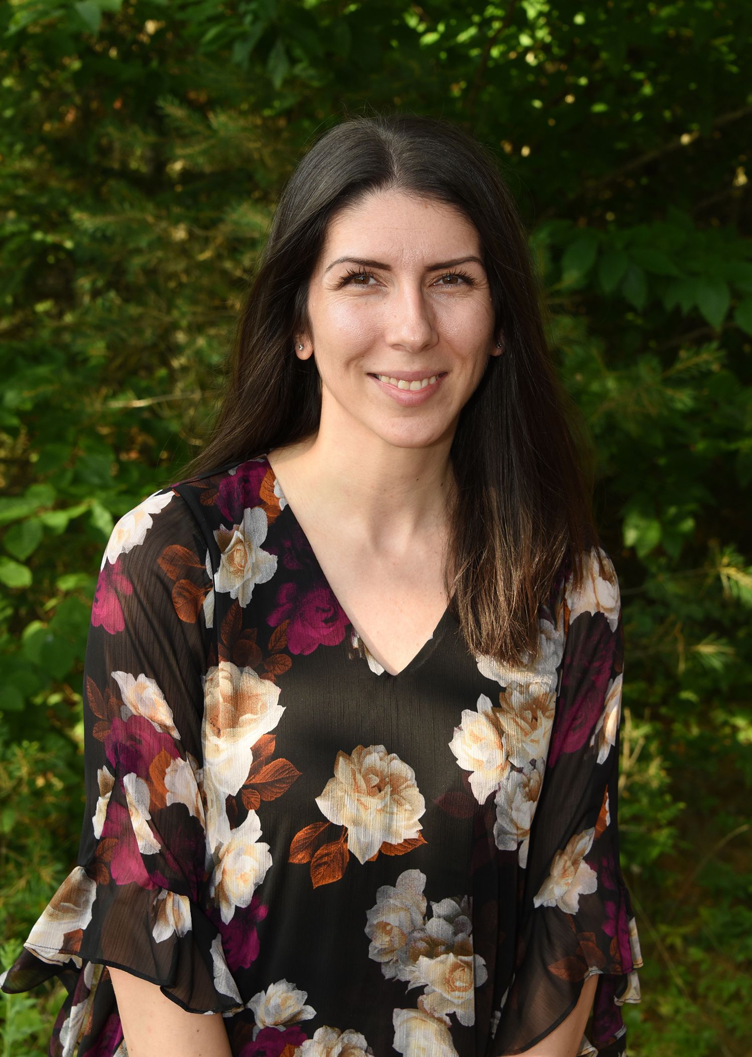 Woman with long dark hair smiles, wearing a black floral print top, standing outdoors with a blurred green background.