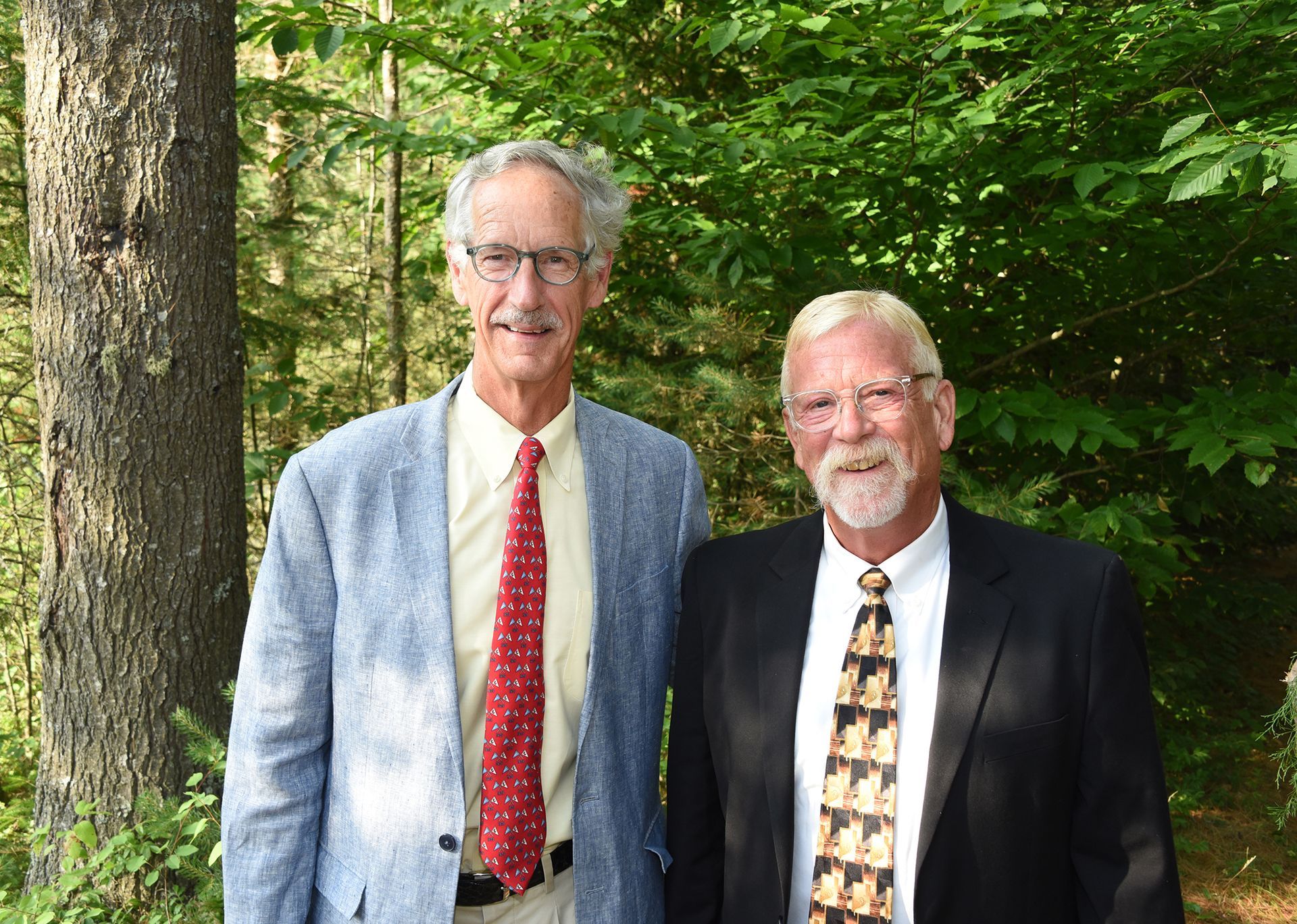 Two men standing outdoors; one in a blue jacket, the other in a black suit. Both are smiling.