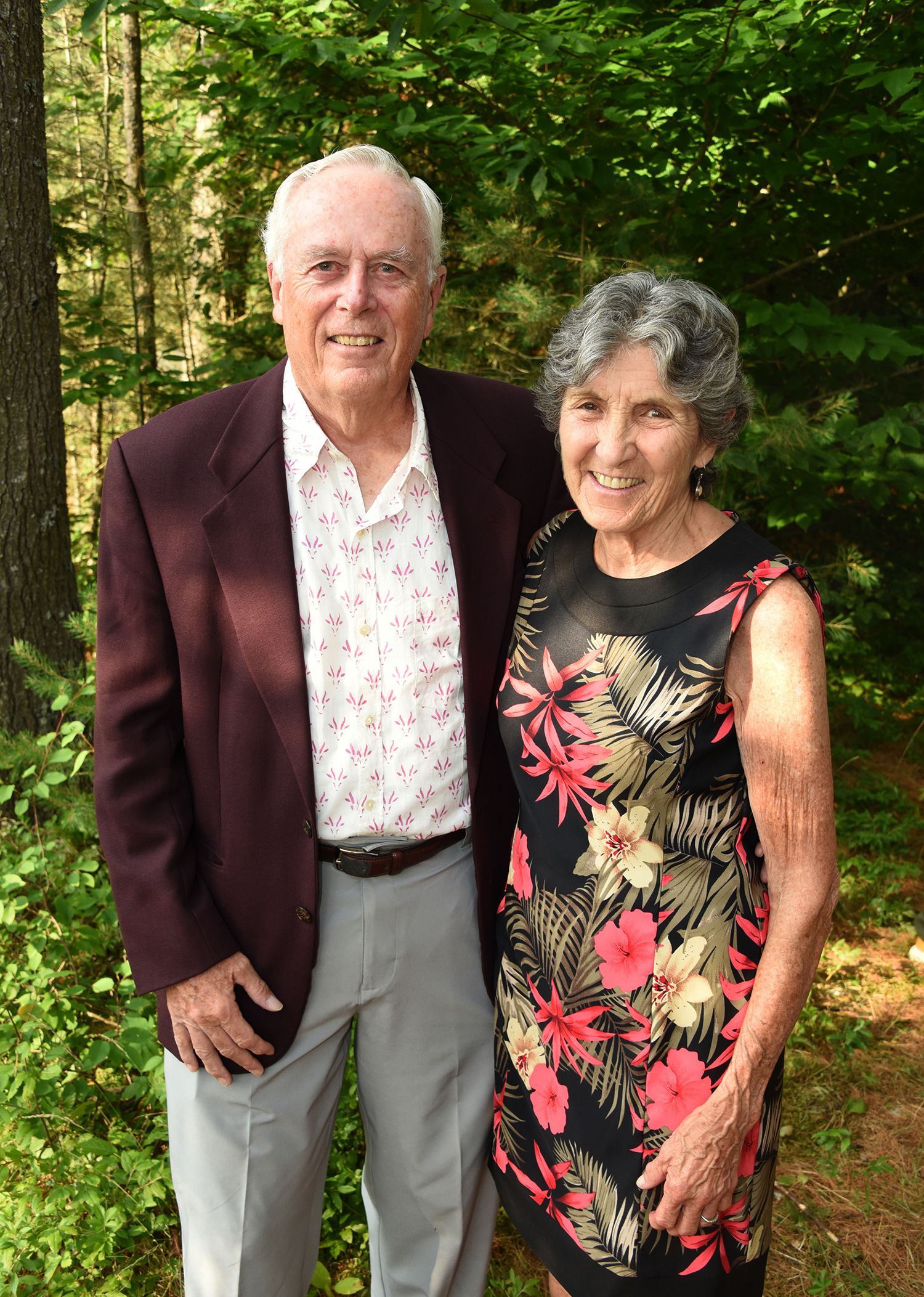 An elderly couple stands in a wooded area. The man wears a maroon blazer and the woman wears a floral dress