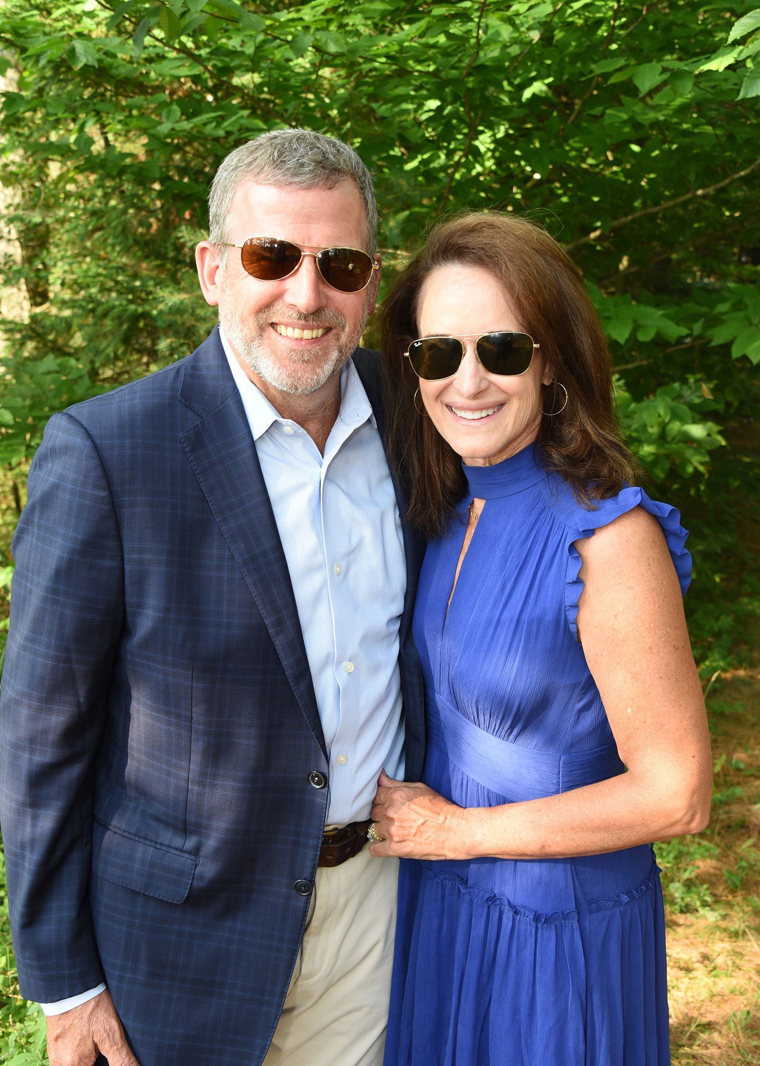 A smiling man and woman pose outdoors. The man wears a navy blazer, and the woman wears a blue dress; both wear sunglasses.