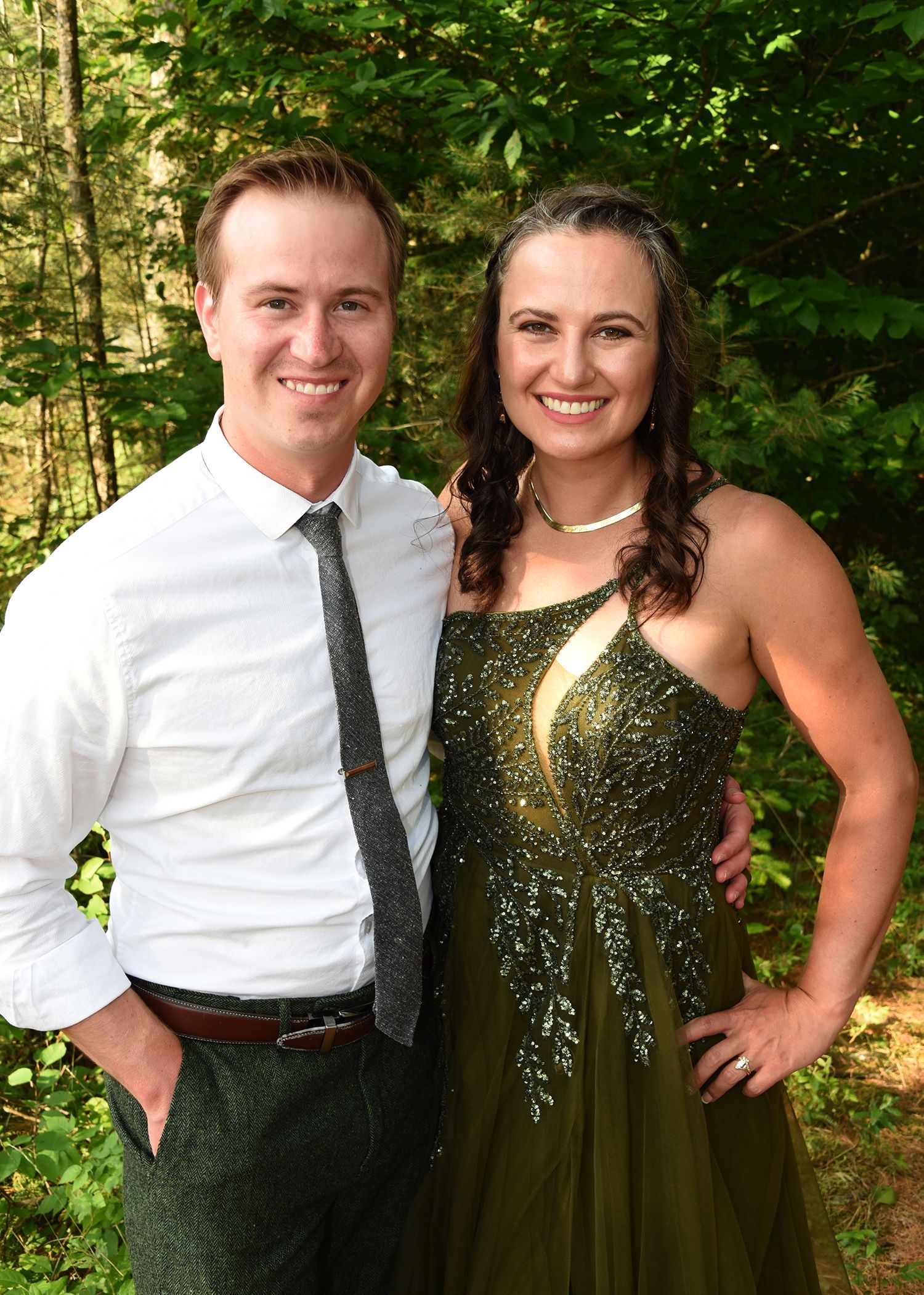 A man and woman pose together outdoors. The man wears a white shirt and tie, the woman a green dress.