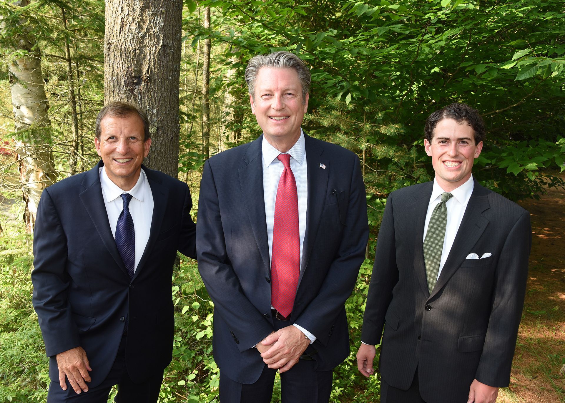 Three men in suits standing outdoors; the man in the middle has a red tie. They are smiling in front of a wooded background.