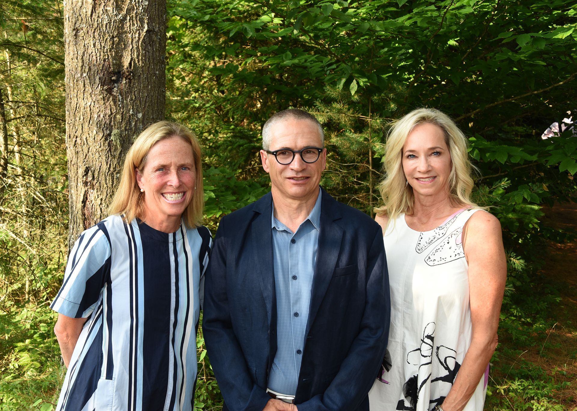Three people pose outdoors in front of trees. A man in a blue blazer is flanked by two women