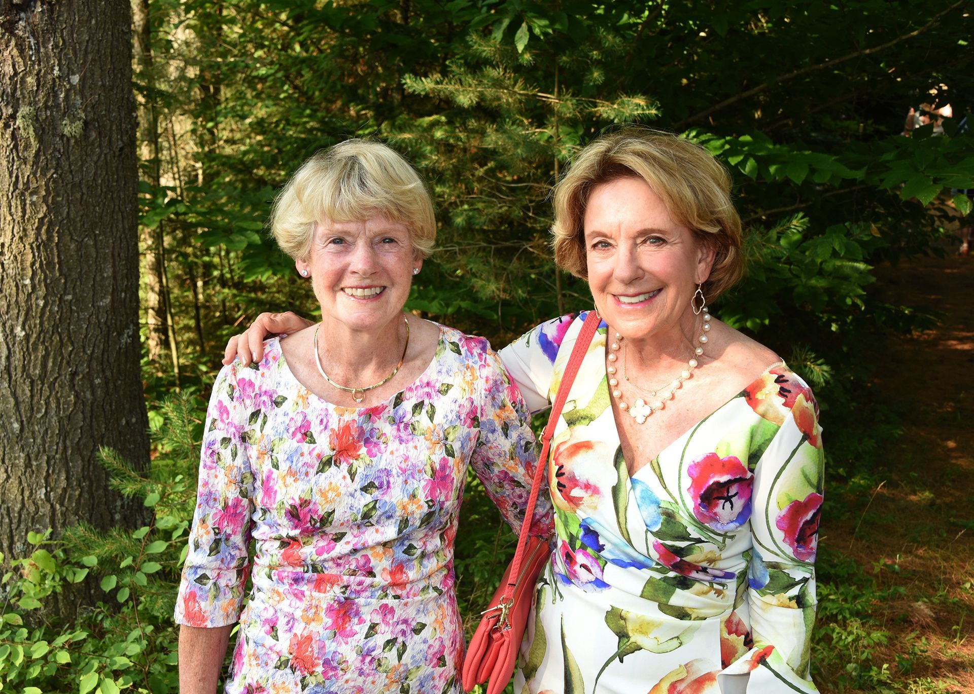 Two women with blonde hair smile and pose outdoors. They wear floral print dresses, standing in front of a tree and greenery.