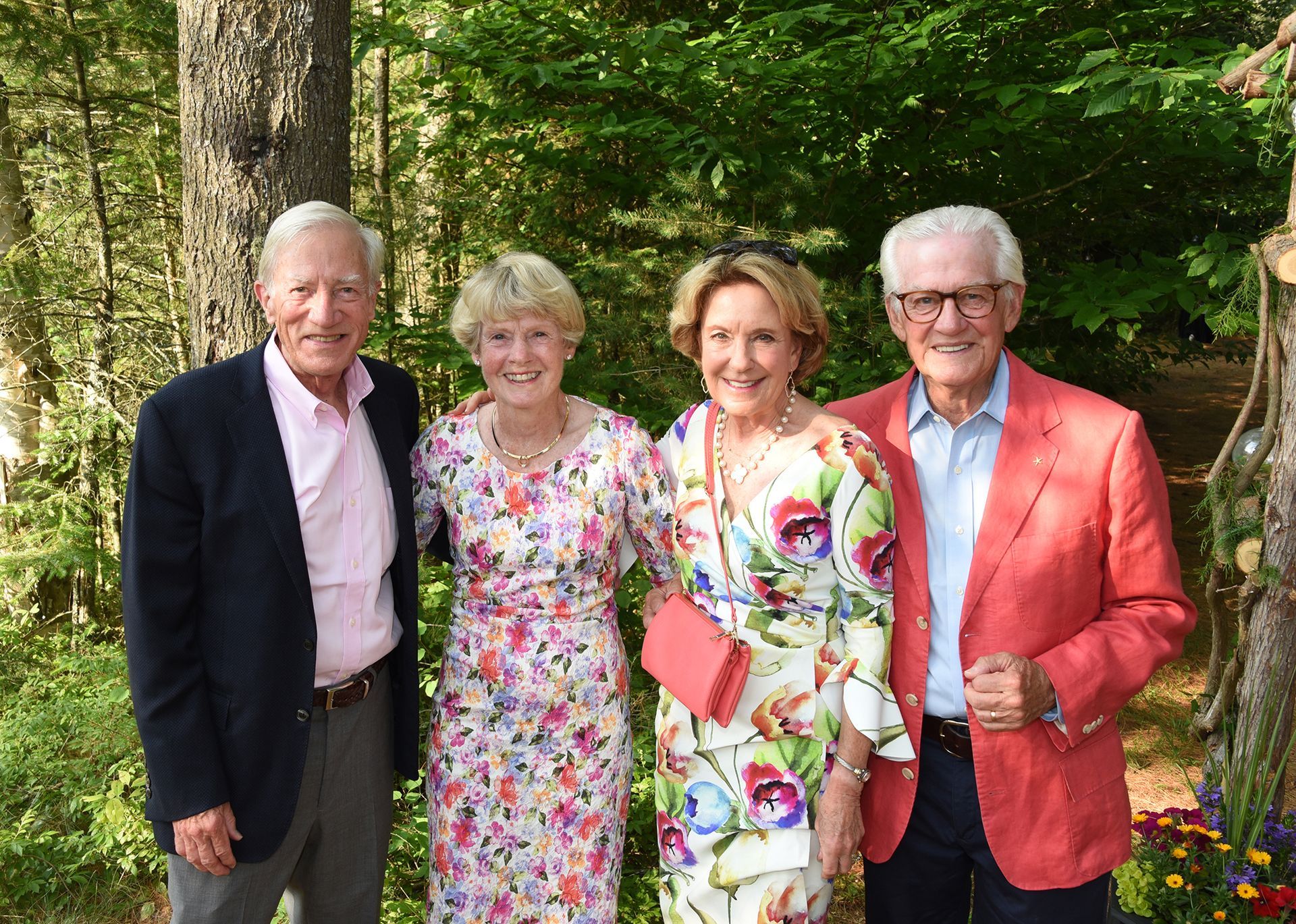 Four well-dressed people smiling outdoors. Two women in floral dresses flank two men in jackets, one navy and one coral