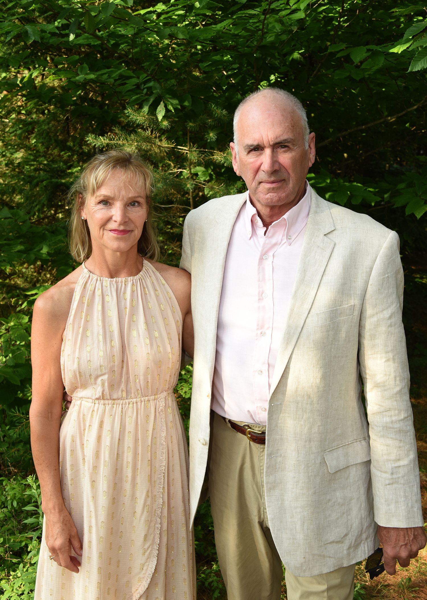 A man and woman stand outdoors. The woman wears a light-colored dress, the man a beige jacket and pink shirt