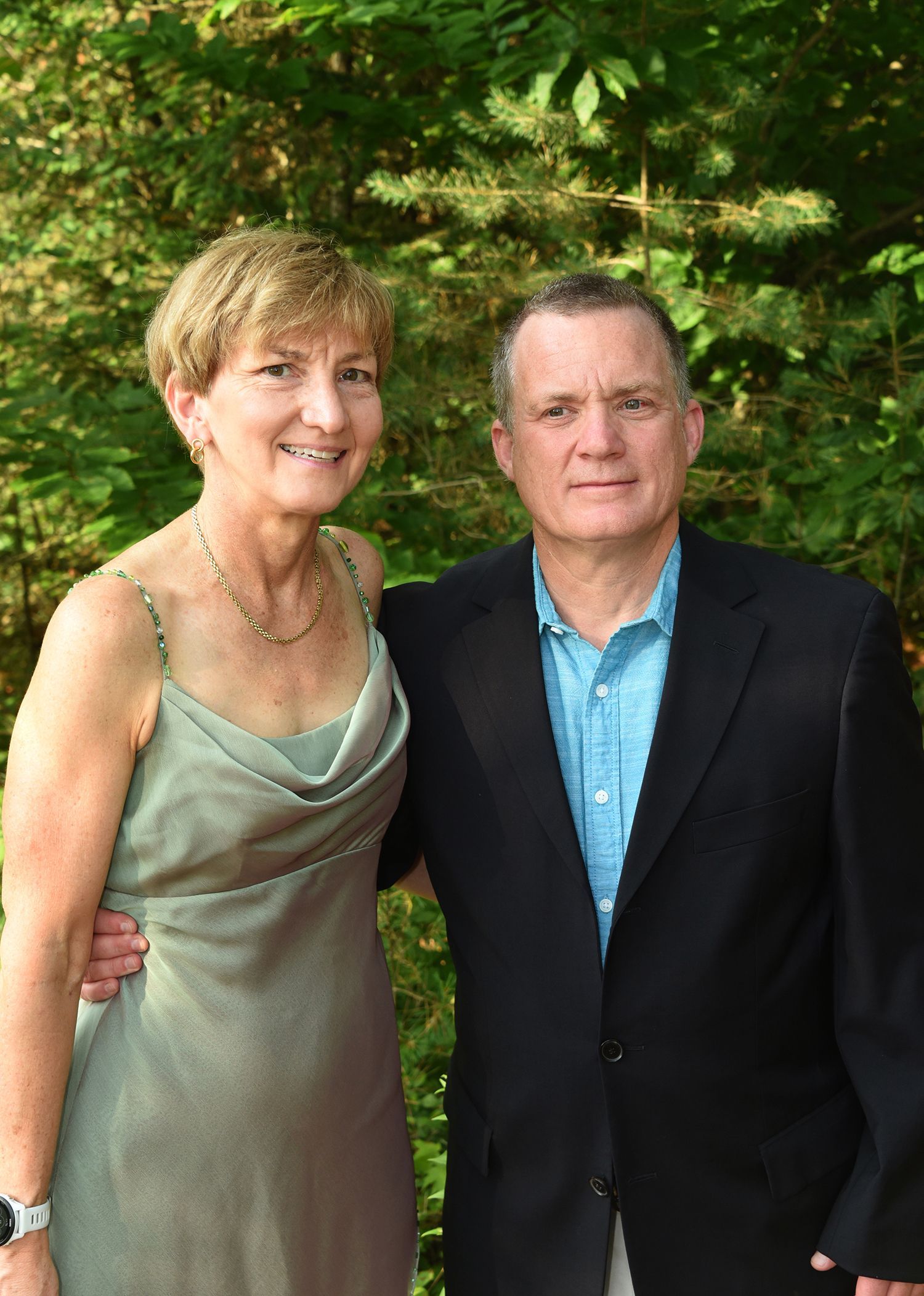 A smiling older woman in a green dress and a man in a blazer and blue shirt pose outdoors, arm in arm.
