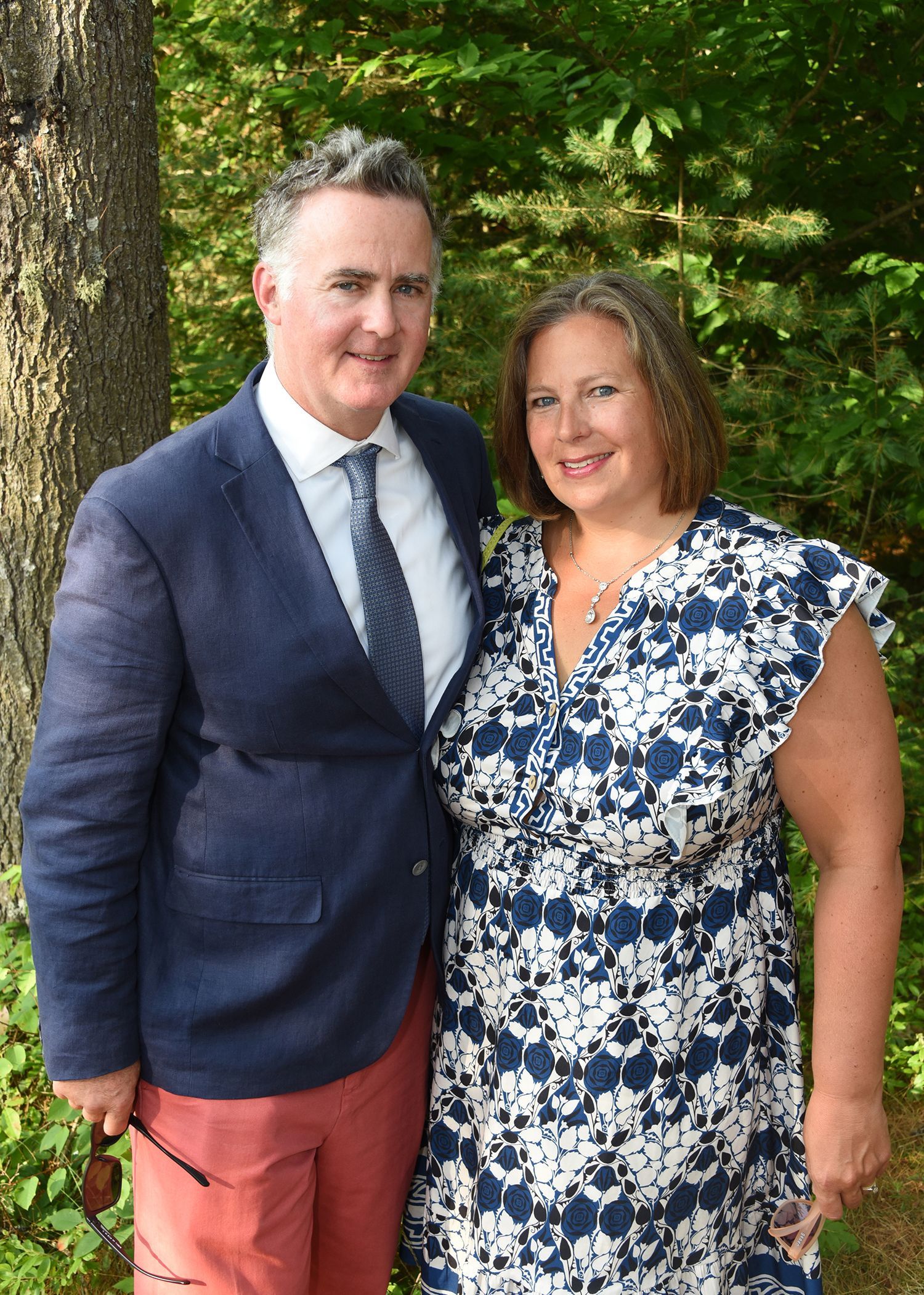 Man and woman standing outdoors. Man in navy jacket and red pants; woman in blue floral dress. They are smiling.