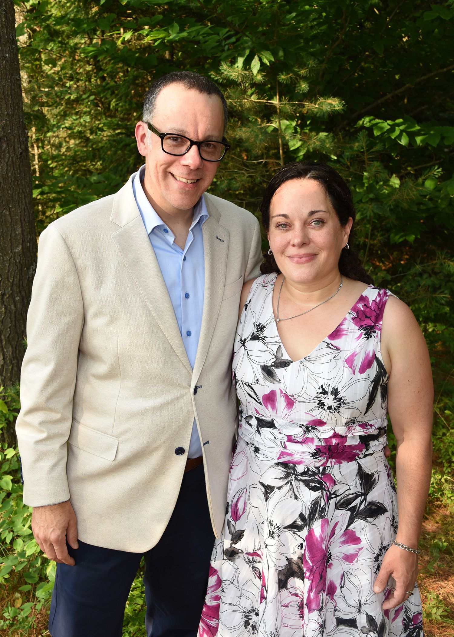A smiling man and woman pose together outdoors. The man wears glasses, a light blazer, and a blue shirt.