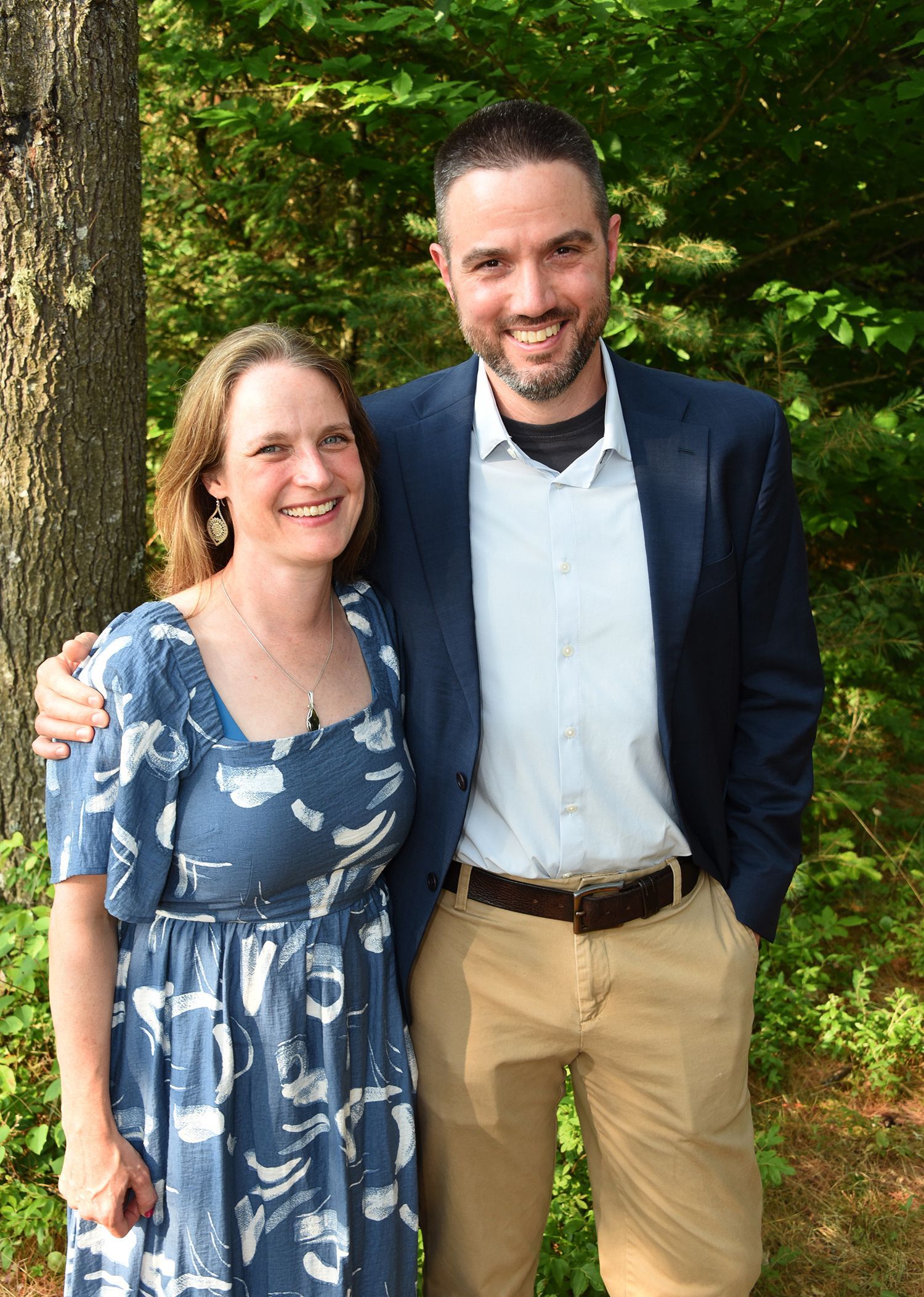 Smiling couple poses outdoors; woman in blue dress, man in blazer and khakis. They are standing next to a tree.