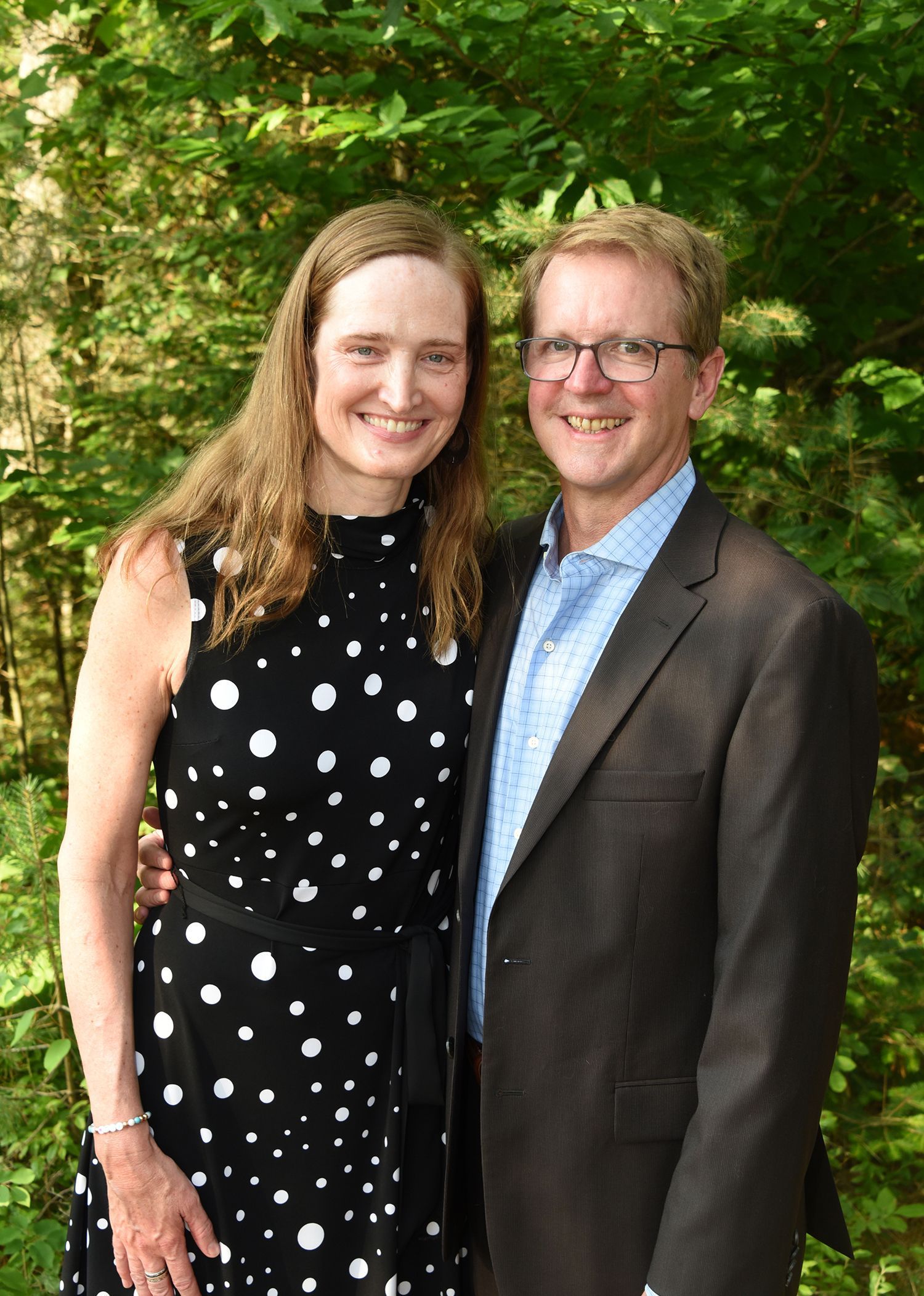 Woman and man smiling, arm-in-arm, in front of green foliage. 