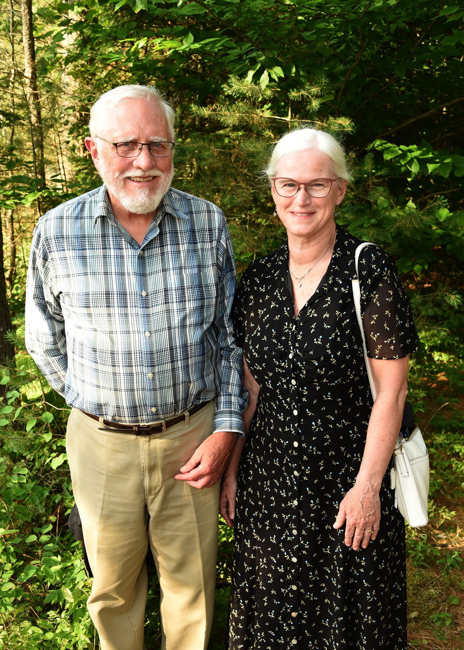 An older man and woman stand outside. The man wears a plaid shirt and khakis, the woman a black dress, both wear glasses.