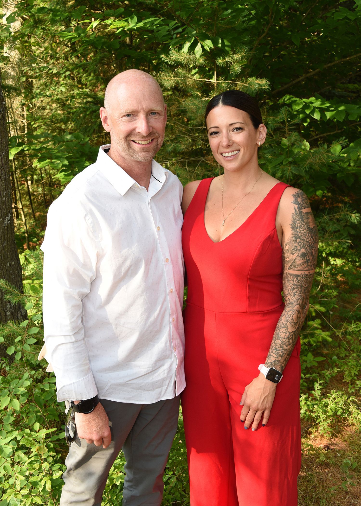 A man and woman pose together outdoors. The man wears a white shirt and grey pants. The woman wears a red jumpsuit.