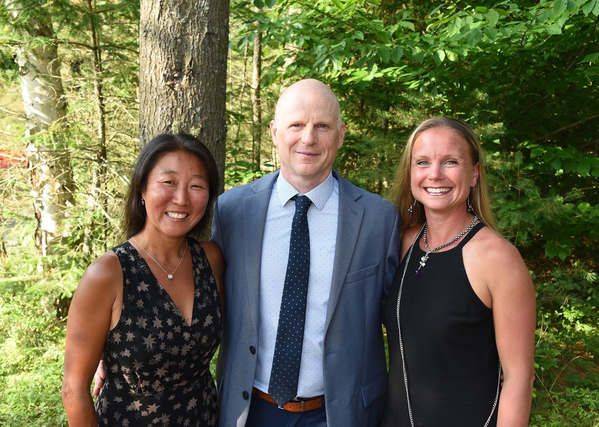 Three smiling people pose outdoors: a woman in a black floral dress, a bald man in a blue suit, and a woman in a black dress.