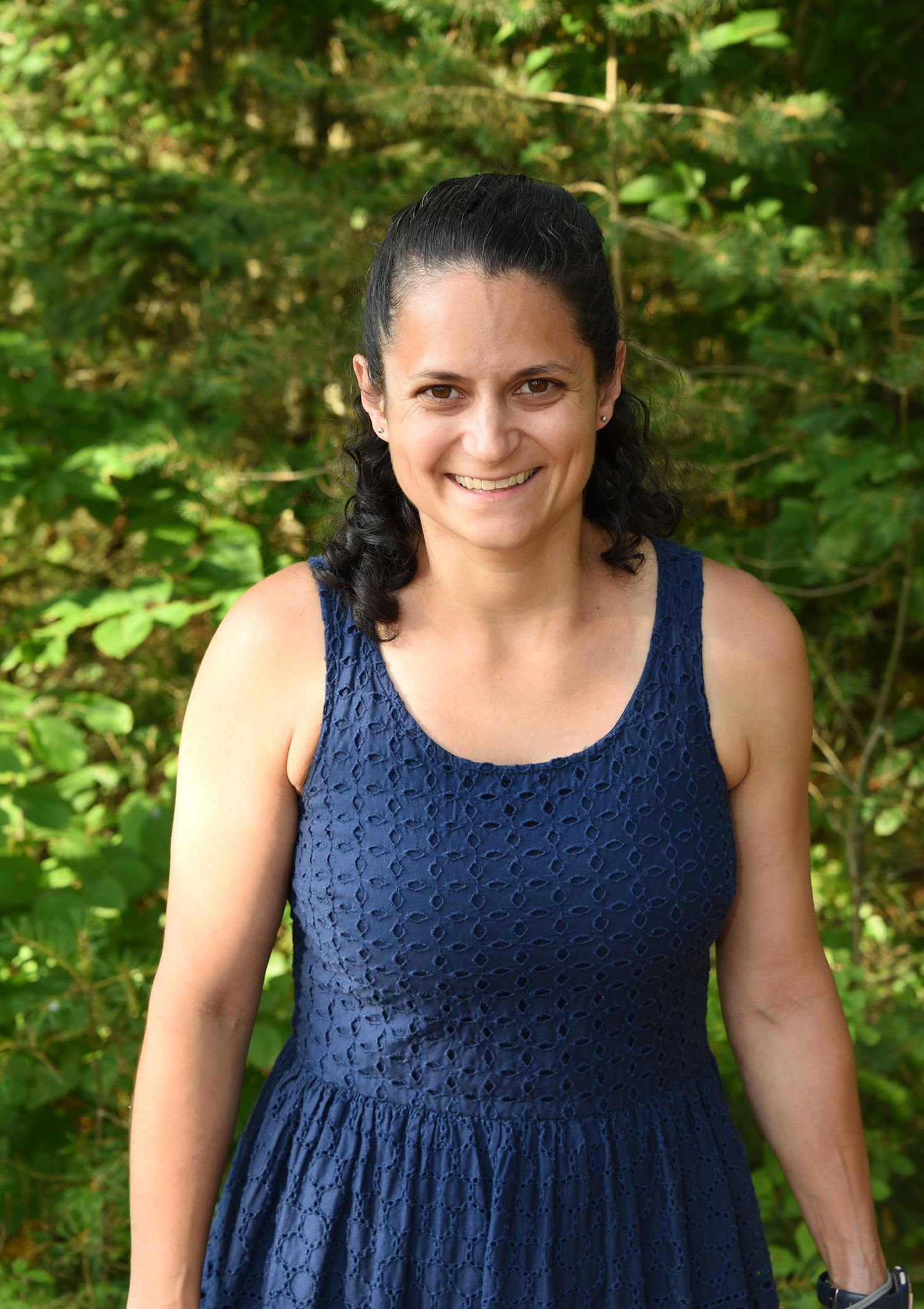 Woman with dark hair and a blue dress smiles outside.