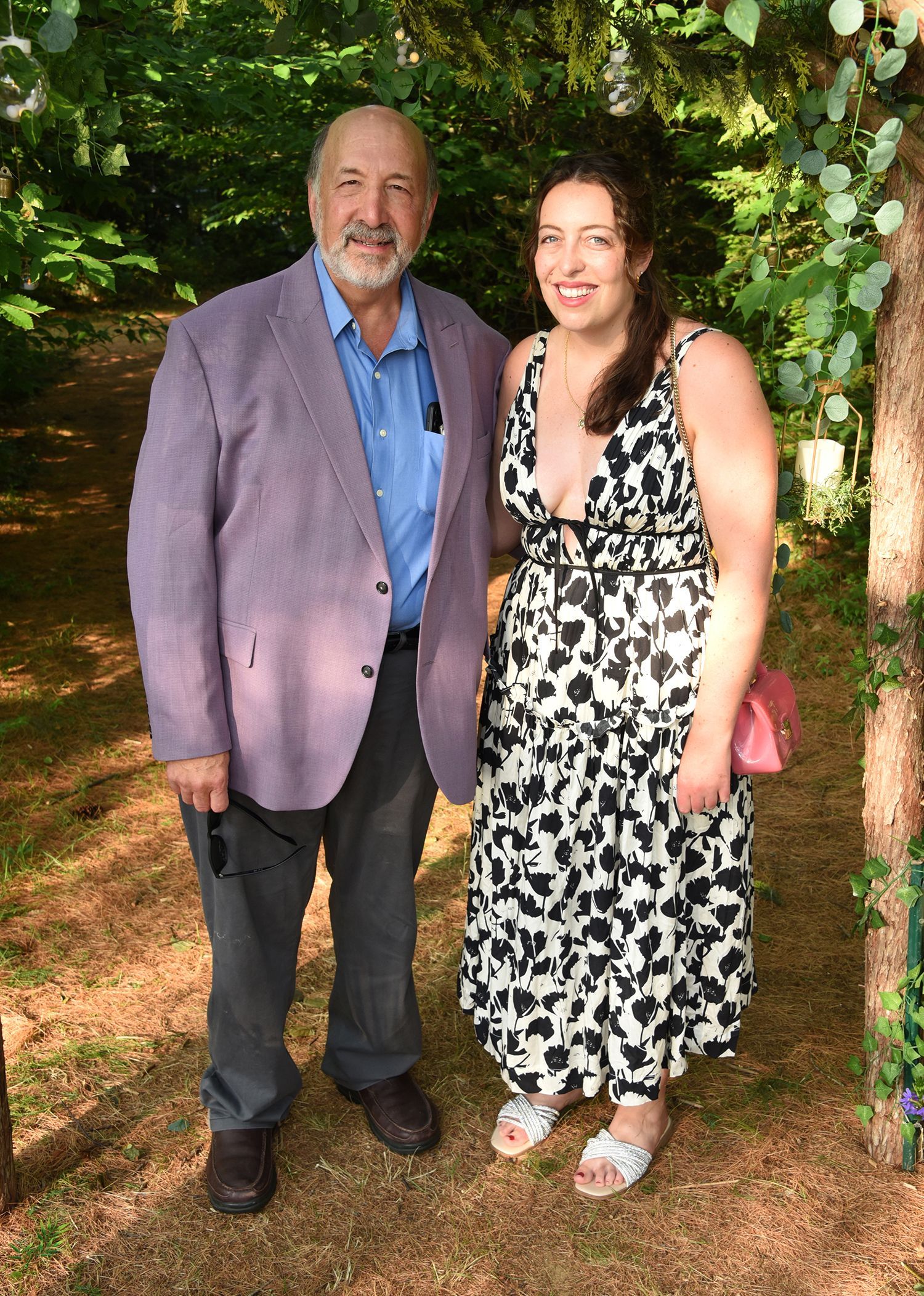 Man and woman standing together outdoors. Man in a purple blazer, woman in a black and white patterned dress. 