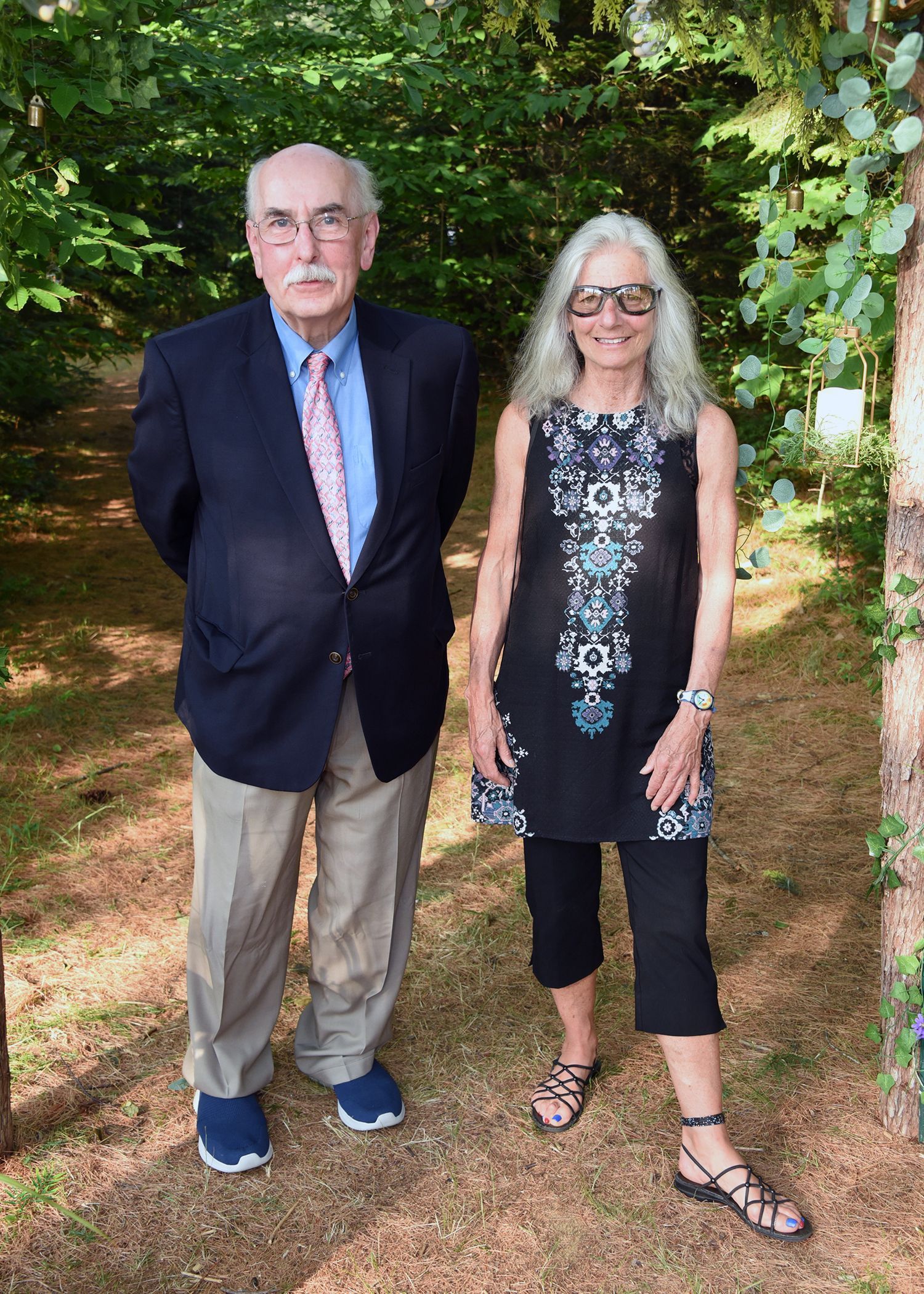 A man and woman pose outdoors. The man wears a navy blazer and khakis. The woman wears a black tunic and capri pants.