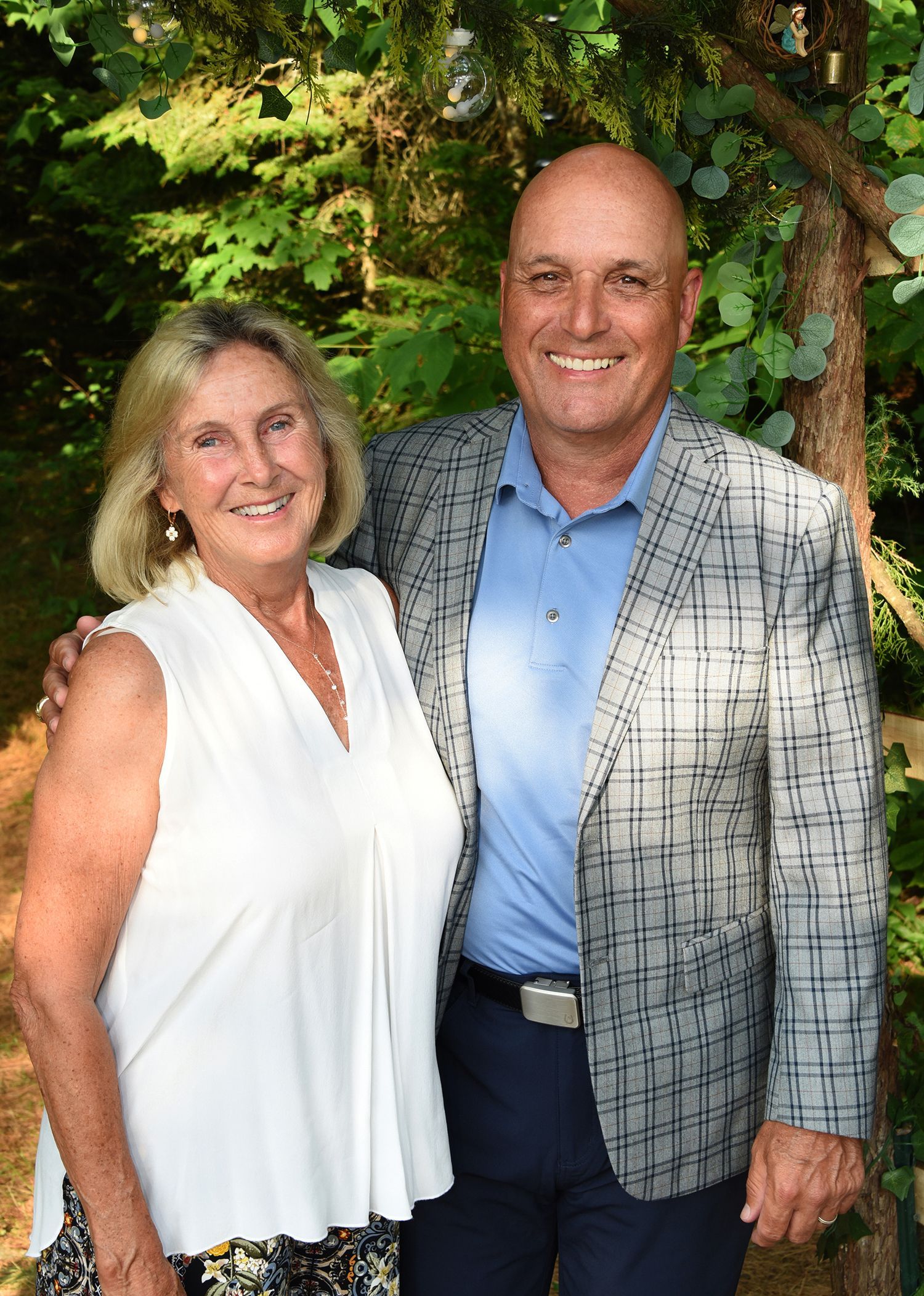 A smiling older couple poses outdoors. The woman wears a white top, the man a blue shirt and patterned blazer.