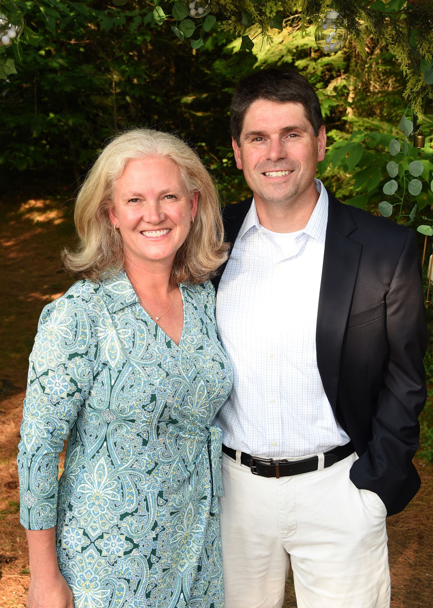 Smiling couple posing outdoors; woman in blue patterned dress, man in blazer, white pants.