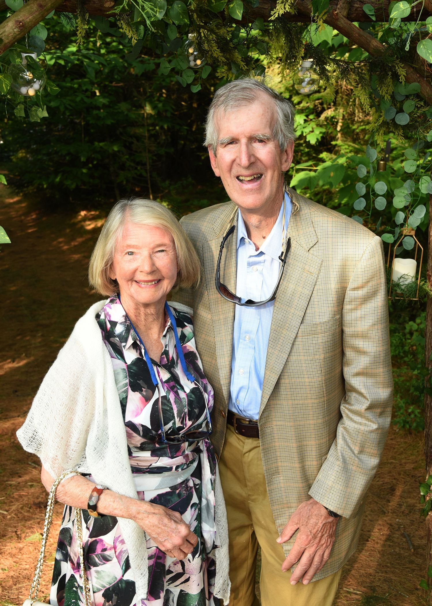 Elderly couple, smiling and posing outdoors. Woman in patterned dress and shawl, man in a light-colored jacket.