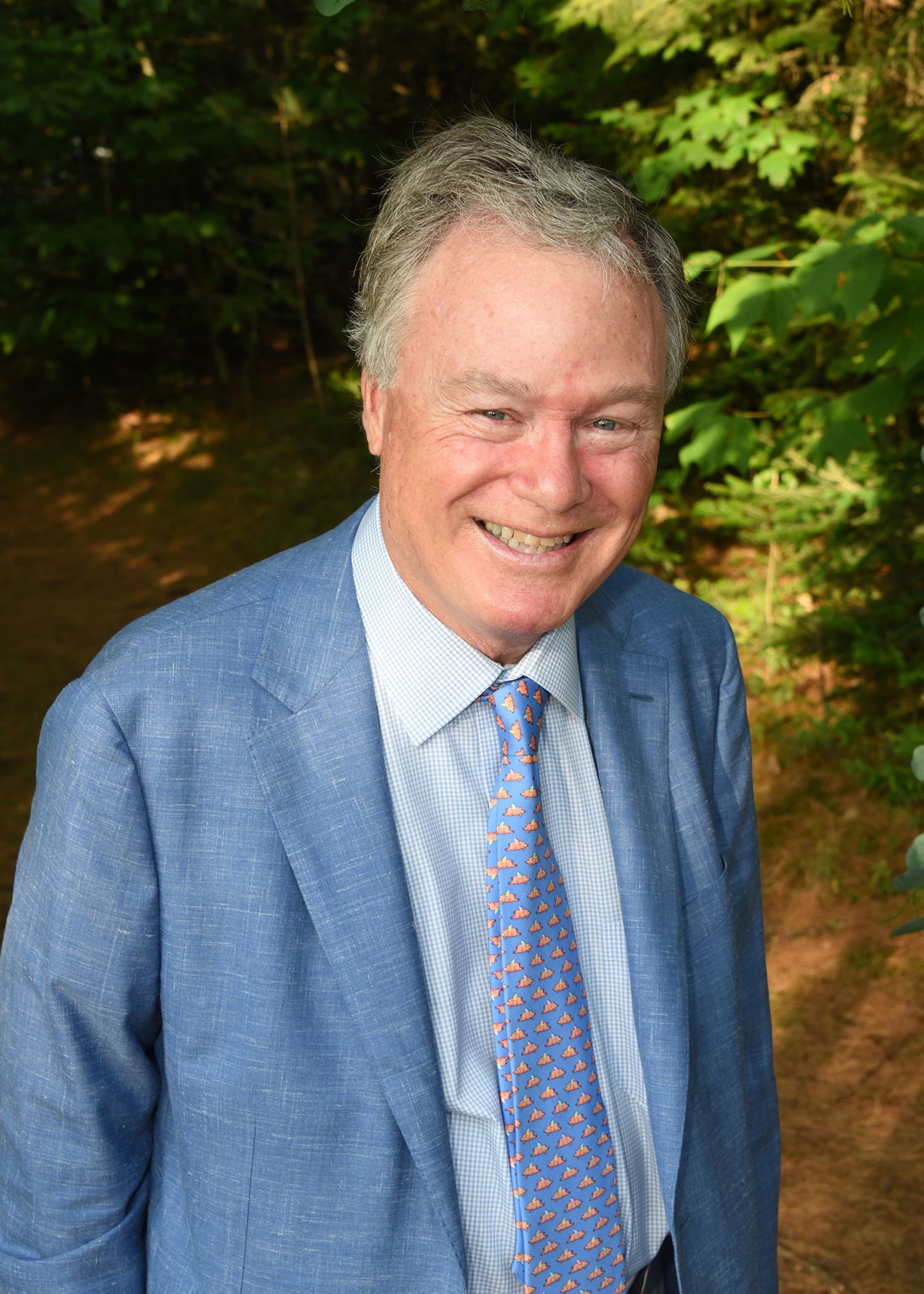 Smiling man in a light blue jacket and patterned tie, outdoors with trees in the background.