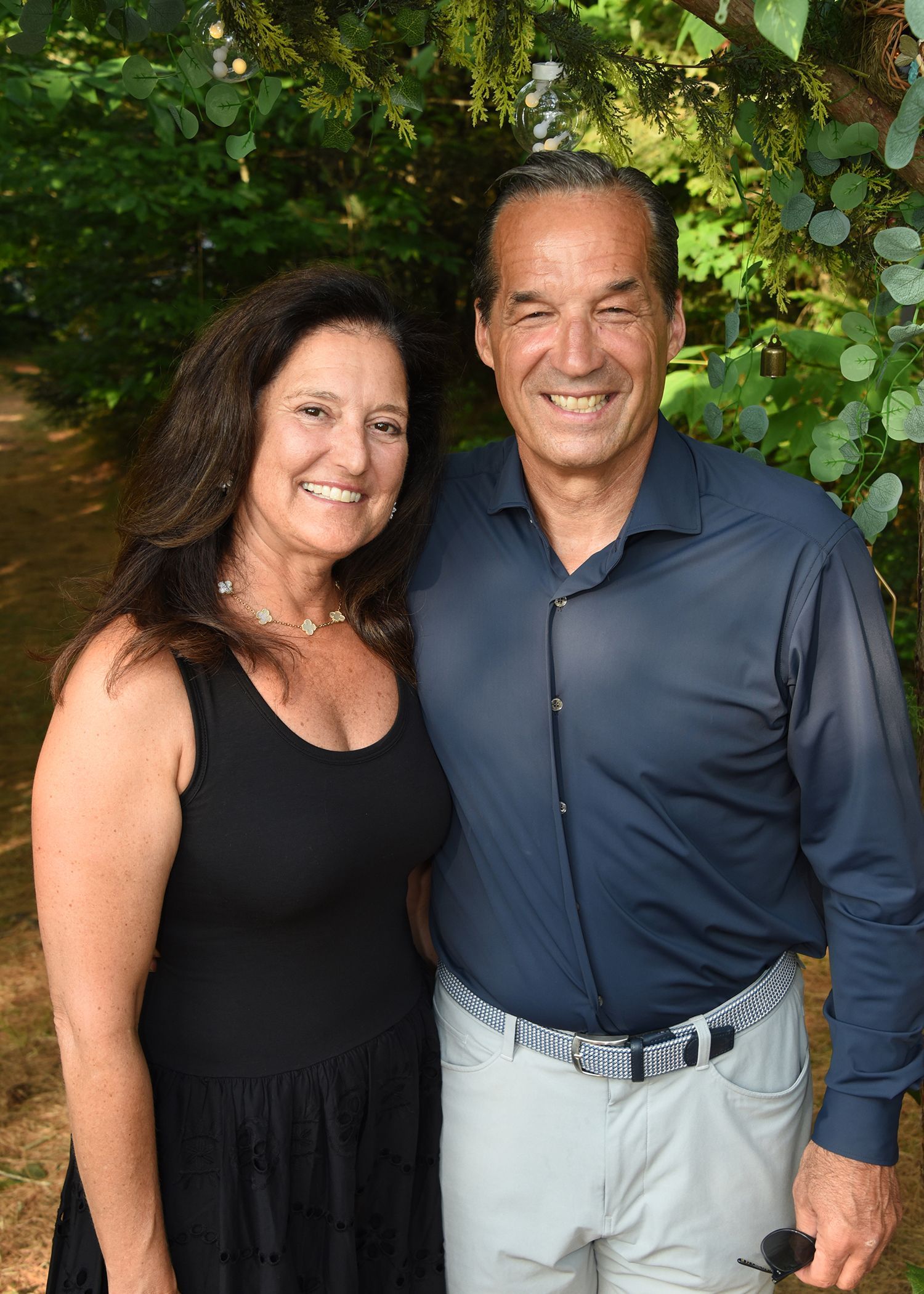 A smiling couple poses outdoors. The woman wears a black dress; the man a blue shirt and light pants.