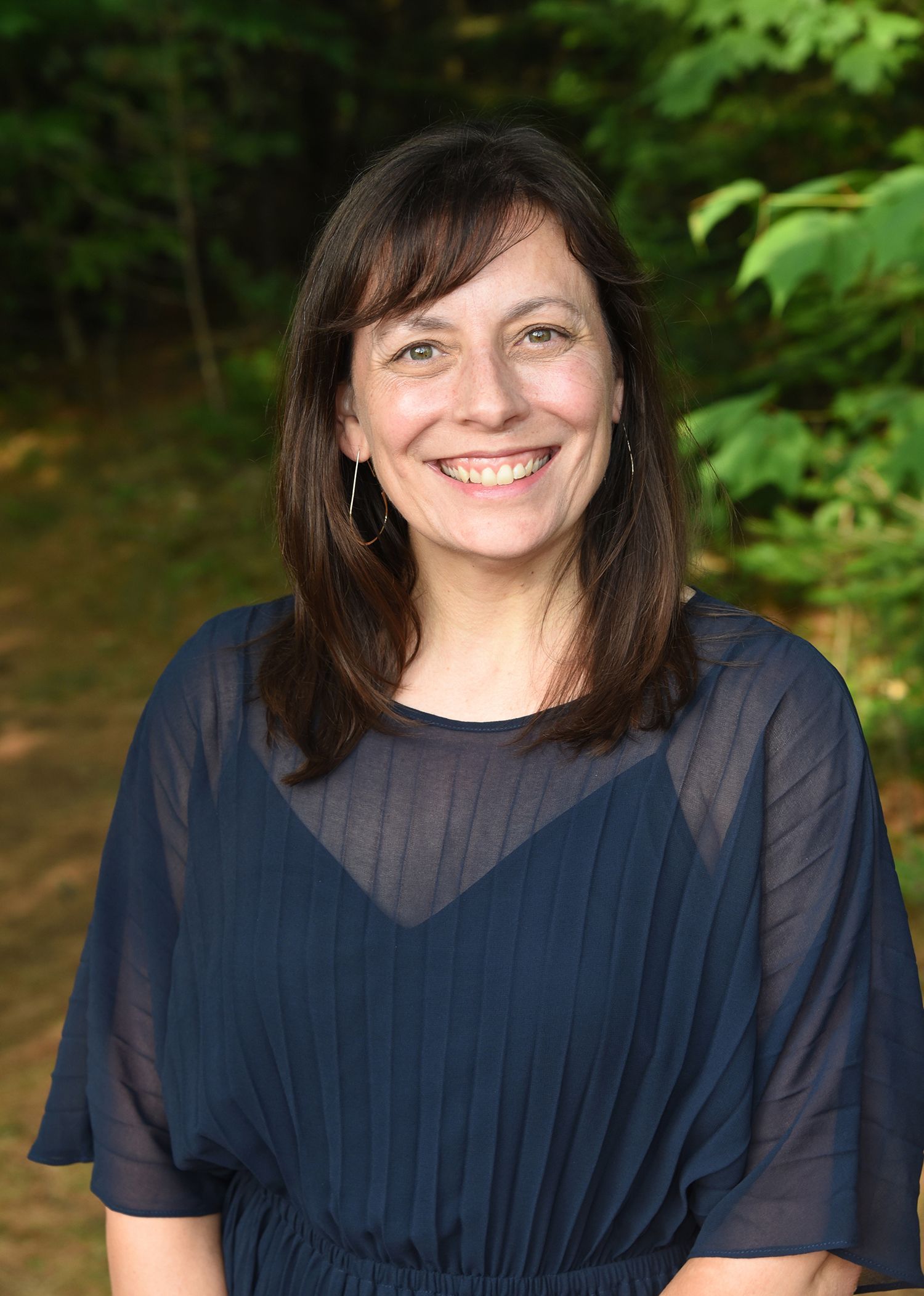 Woman with shoulder-length brown hair smiles outdoors, wearing a sheer blue top over a darker blue garment