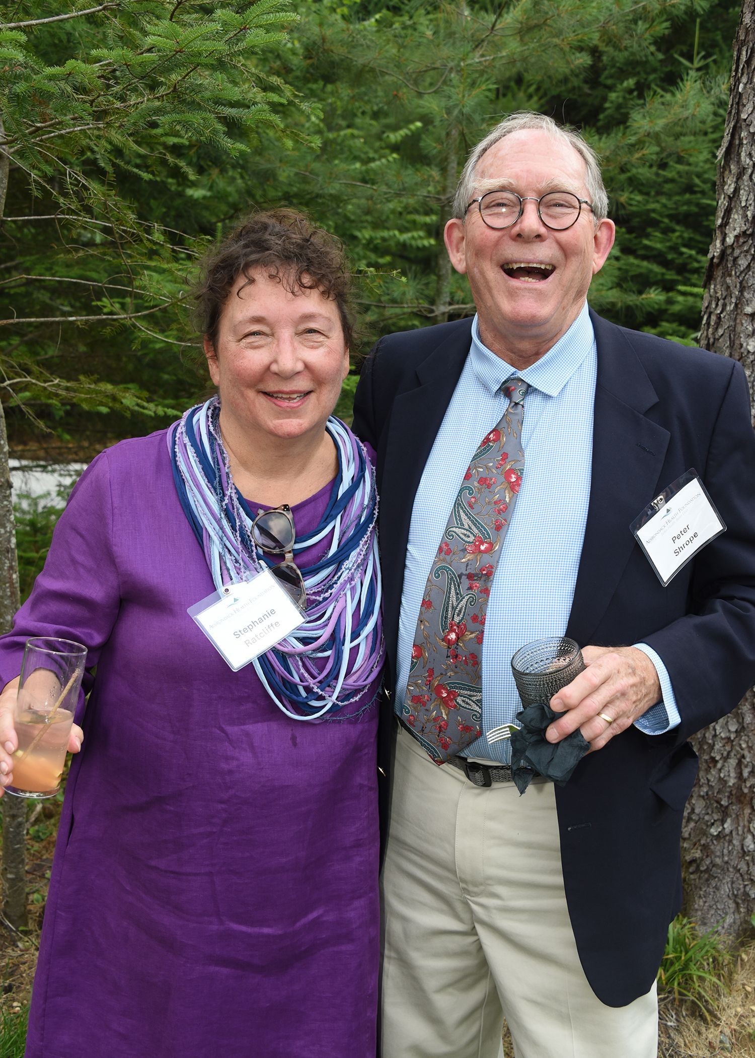 Smiling older couple posing outdoors. Woman in purple dress and scarf holds a drink.