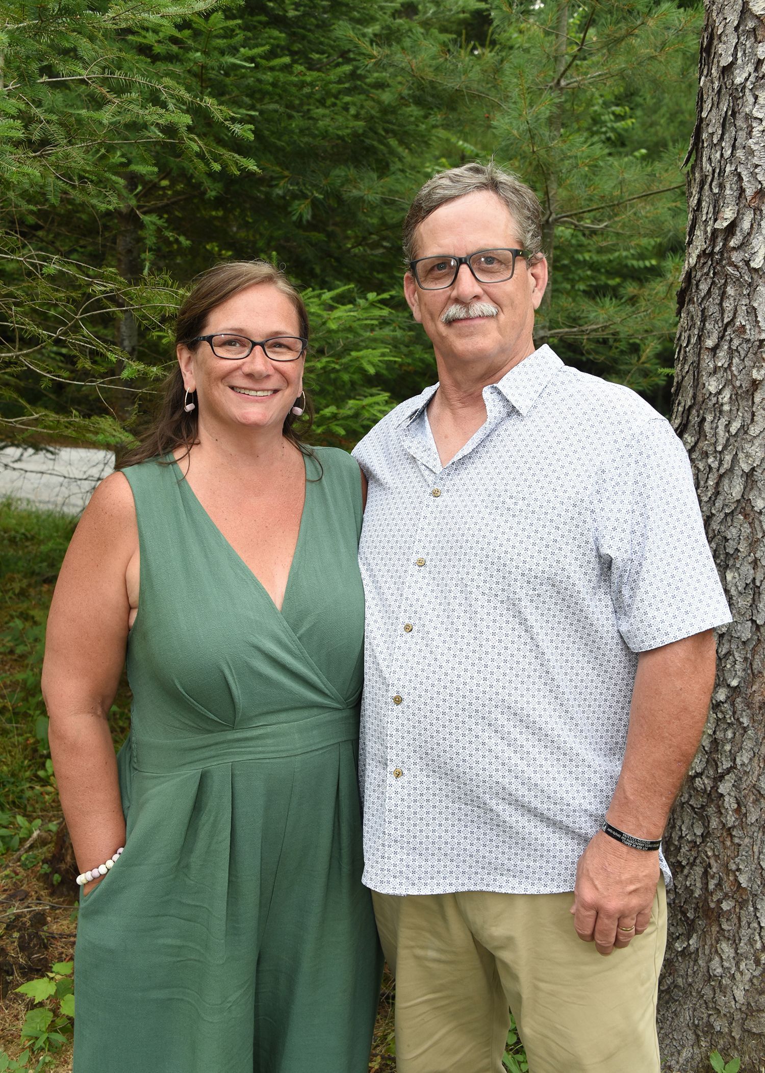 A smiling couple poses outdoors. Woman wears a green dress; man wears a patterned shirt and khaki pants.