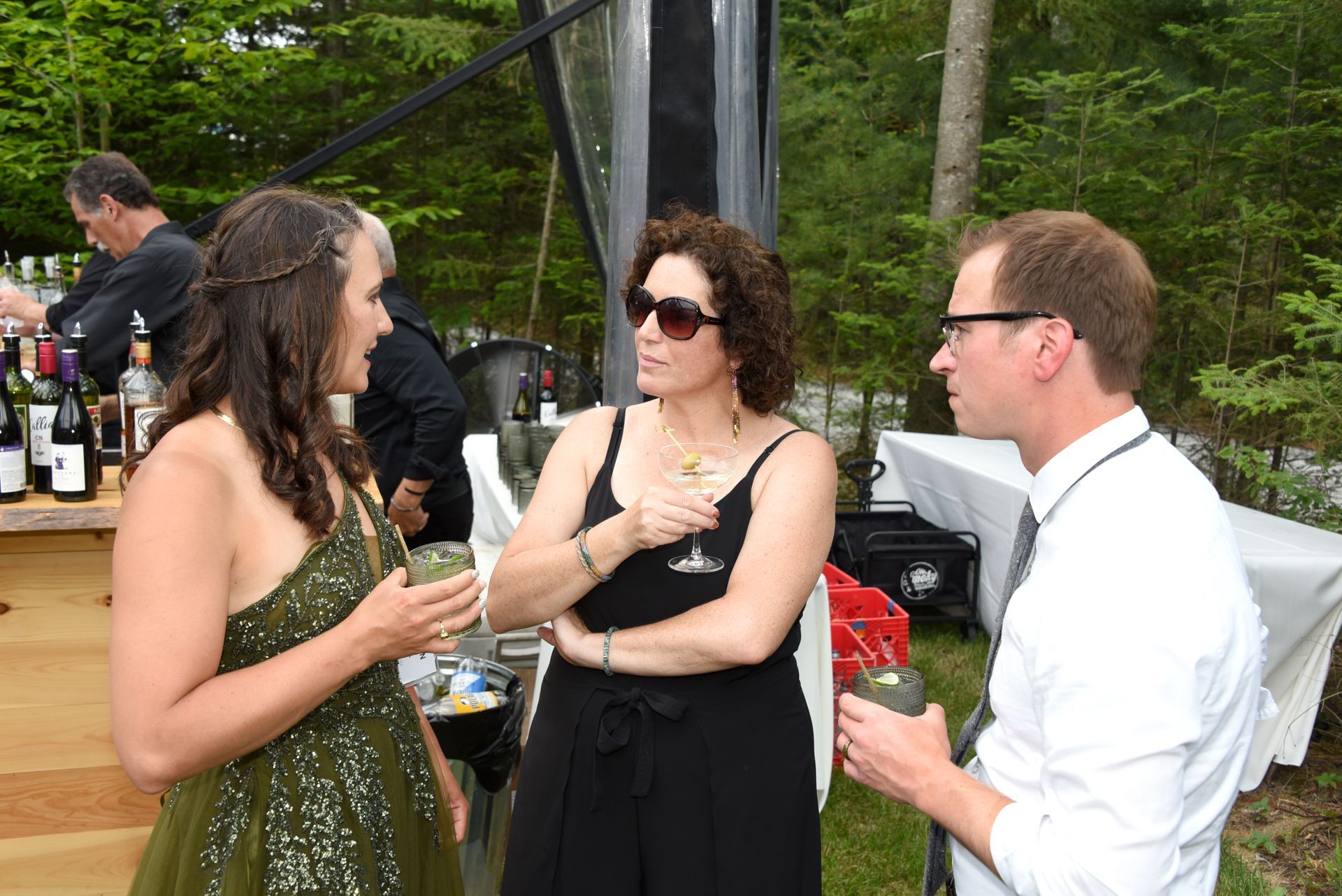 Three people conversing at an outdoor event, near a bar. Woman in green dress, woman in black dress, and man in white shirt.