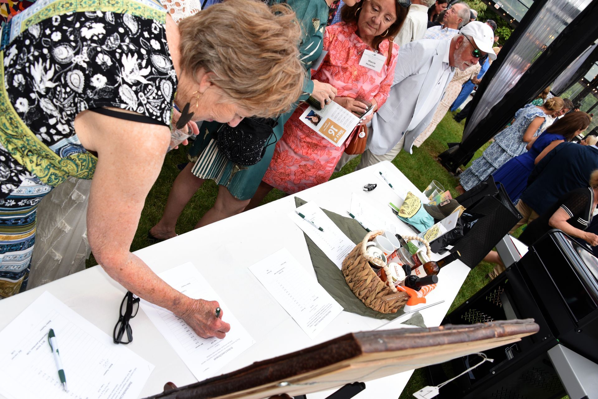 A woman signing a document at a white table, possibly an event registration. Other people stand nearby.