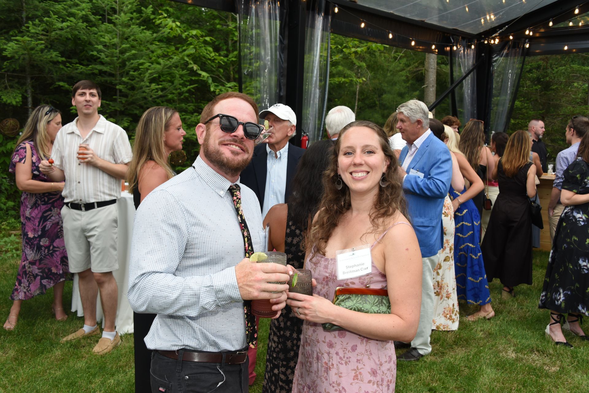 A smiling couple poses at an outdoor party. The man wears sunglasses, a checkered shirt.
