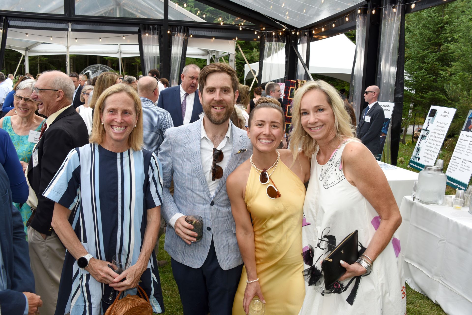 Group of people smiling at an outdoor event; two women in dresses pose with two men in blazers.