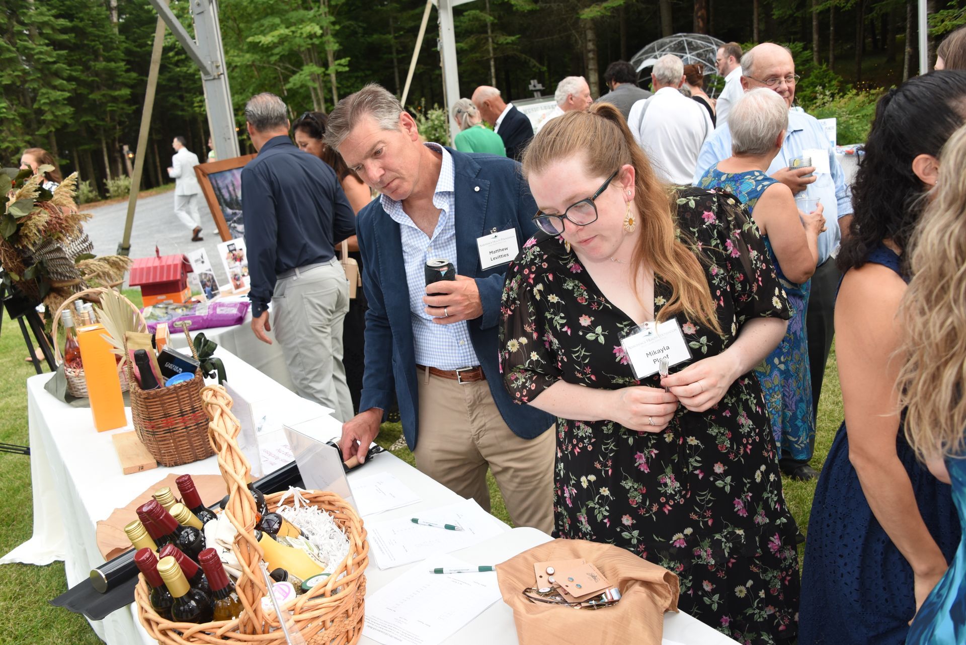 People browsing items at an outdoor event. A man and woman examine items at a table, holding drinks and wearing name tags.