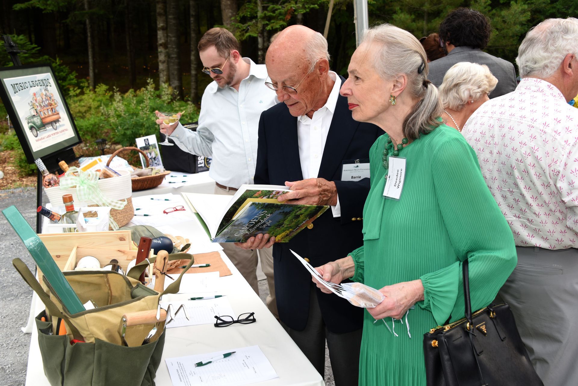 Two people browse a book at an outdoor display table. The woman wears green and the man has a navy blazer. 