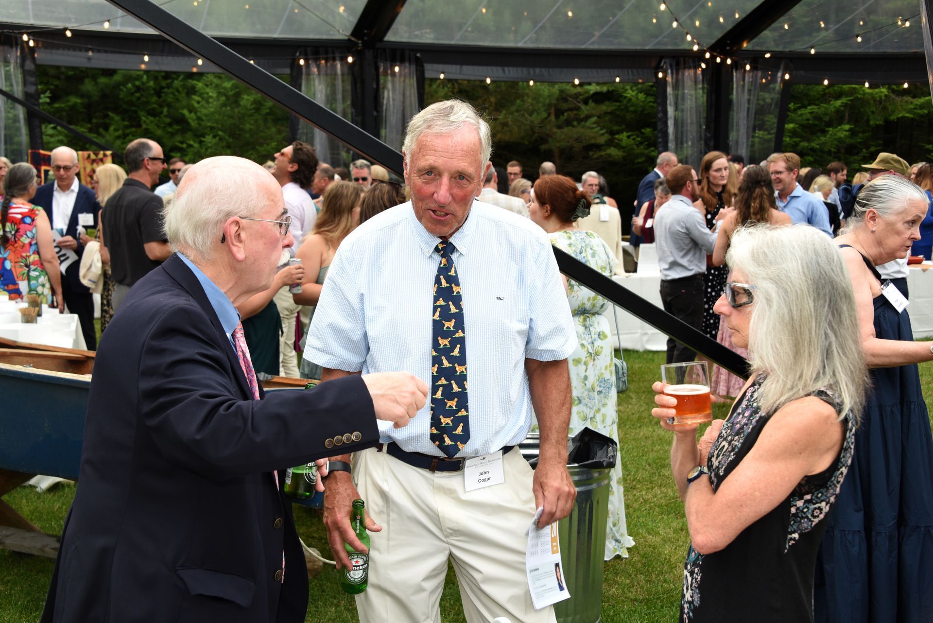Three people chatting at an outdoor event; a man in a blue blazer gestures, another man in a shirt and tie looks on.
