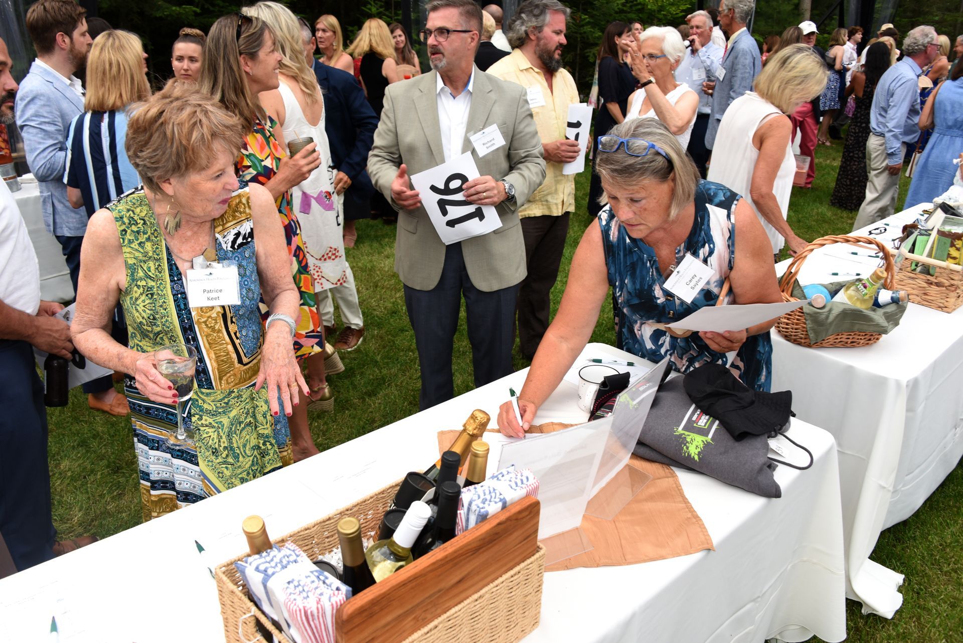 People at an outdoor event with tables of auction items. A woman examines a prize, another looks on. Others mingle.