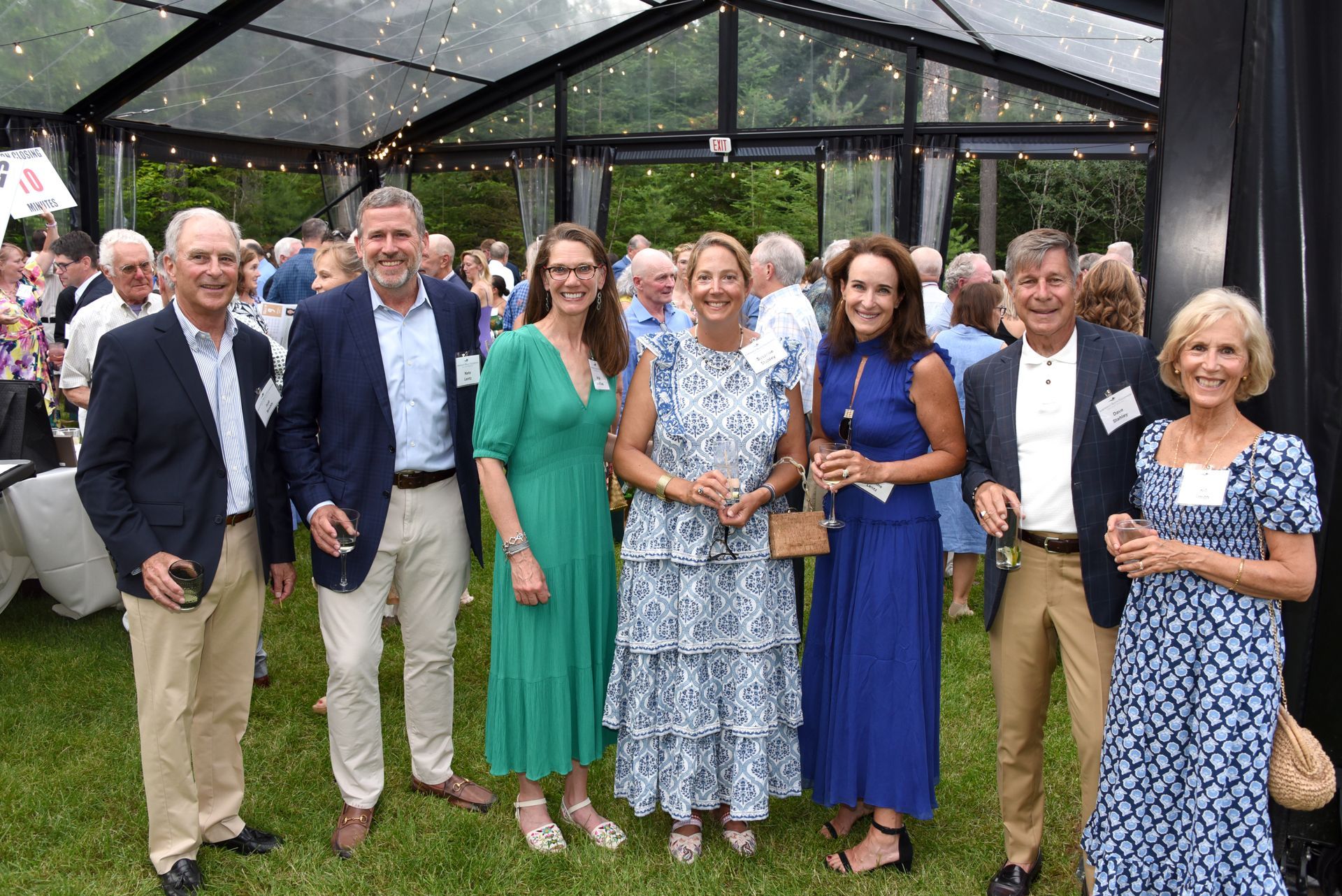 A group of eight people, elegantly dressed, poses for a photo at an outdoor event under a clear tent.