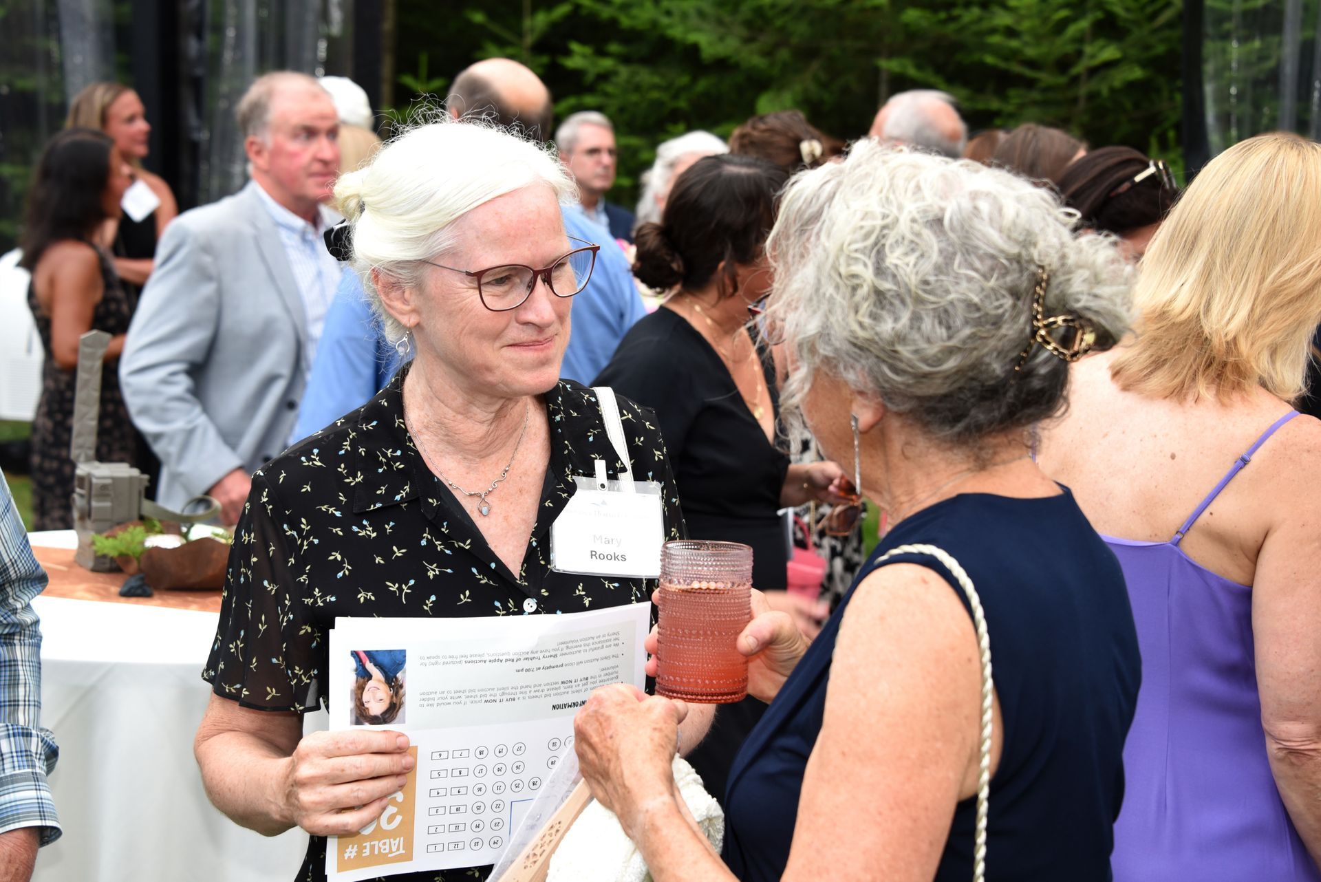Two women chatting at an outdoor gathering. One holds a paper with a photo; the other, a drink. Others mingle nearby.